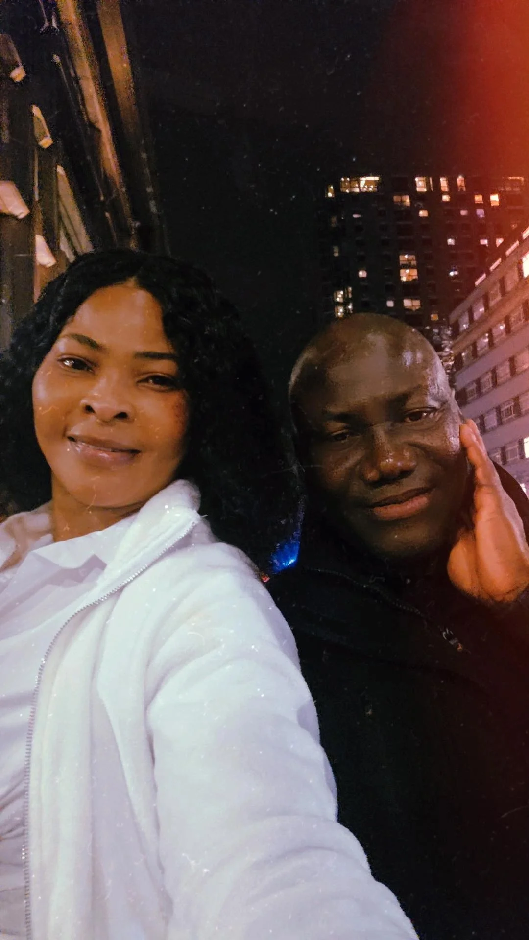 A woman with black curly hair smiling and wearing a white jacket, next to a man with a bald head, resting his hand on his face, both posing for a selfie outdoors at night with city buildings in the background.