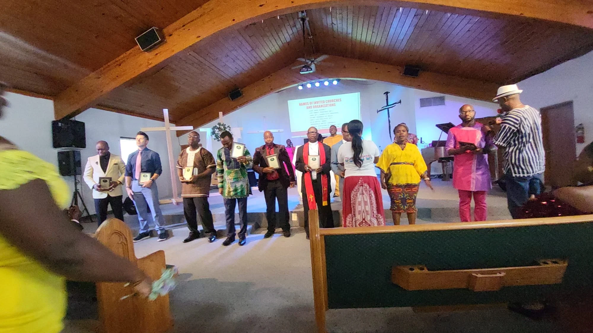 Group of people standing on stage inside a church, some holding plaques or certificates, with a cross on the wall behind them, and a presentation slide projected on the wall.
