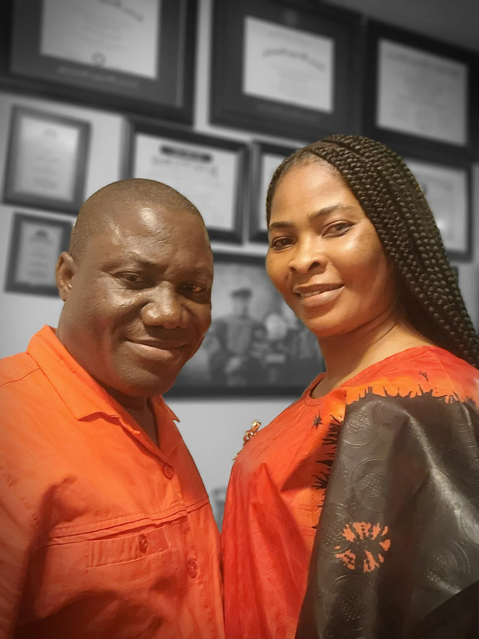 A man and a woman pose for a selfie in front of a wall with framed certificates or awards. The man is wearing an orange shirt, and the woman is dressed in a similar colored outfit with black lace and embroidery details.