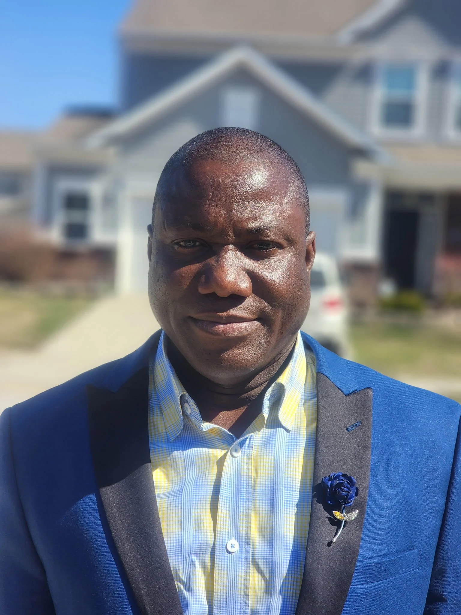 A man in a blue suit and yellow checkered shirt taking a selfie outdoors with a house and clear sky in the background.