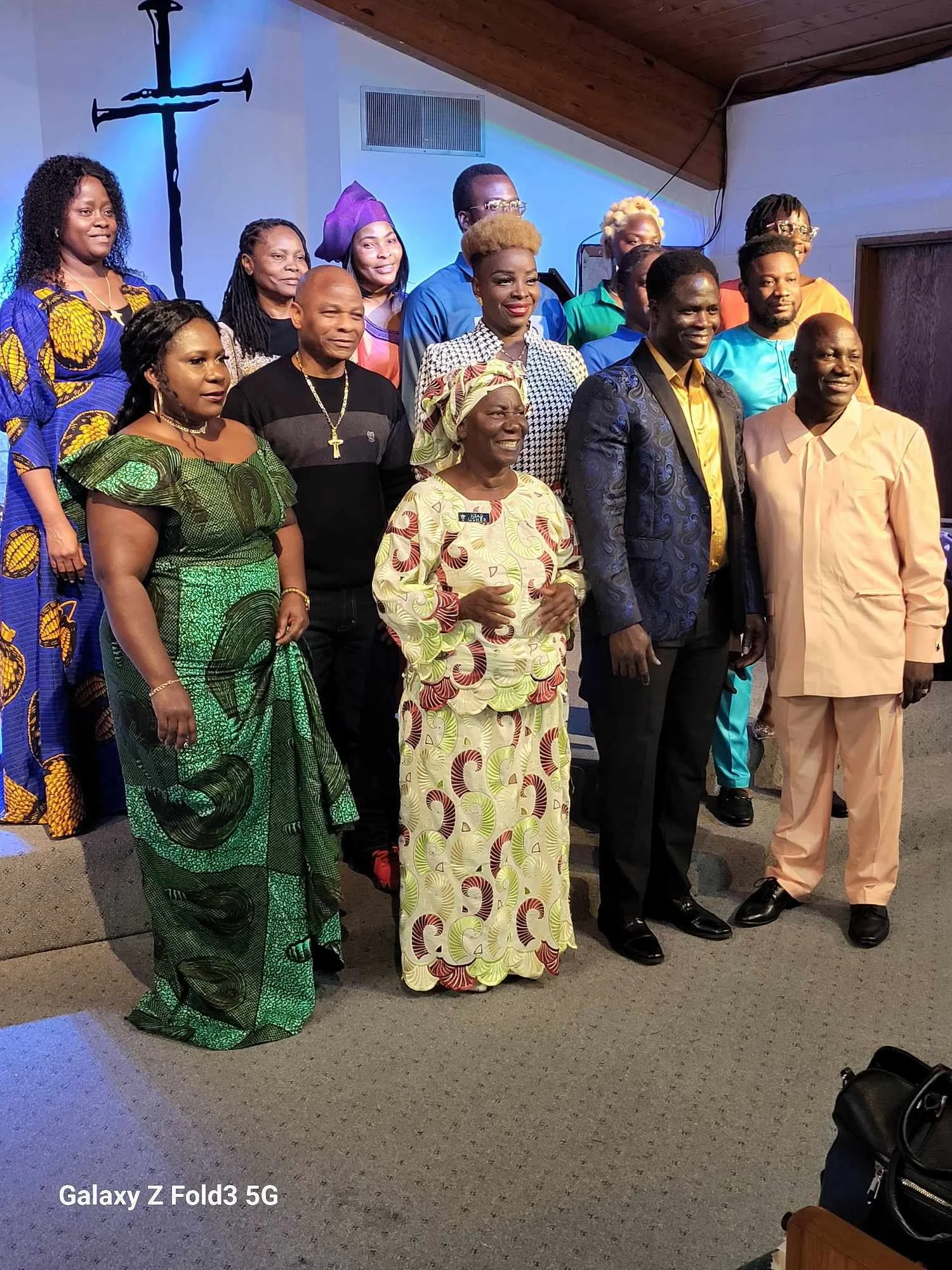 A group of religious leaders and community members posing together indoors in front of a cross on the wall. They are dressed in colorful traditional and formal clothing, standing on a carpeted floor, and smiling for the photo.