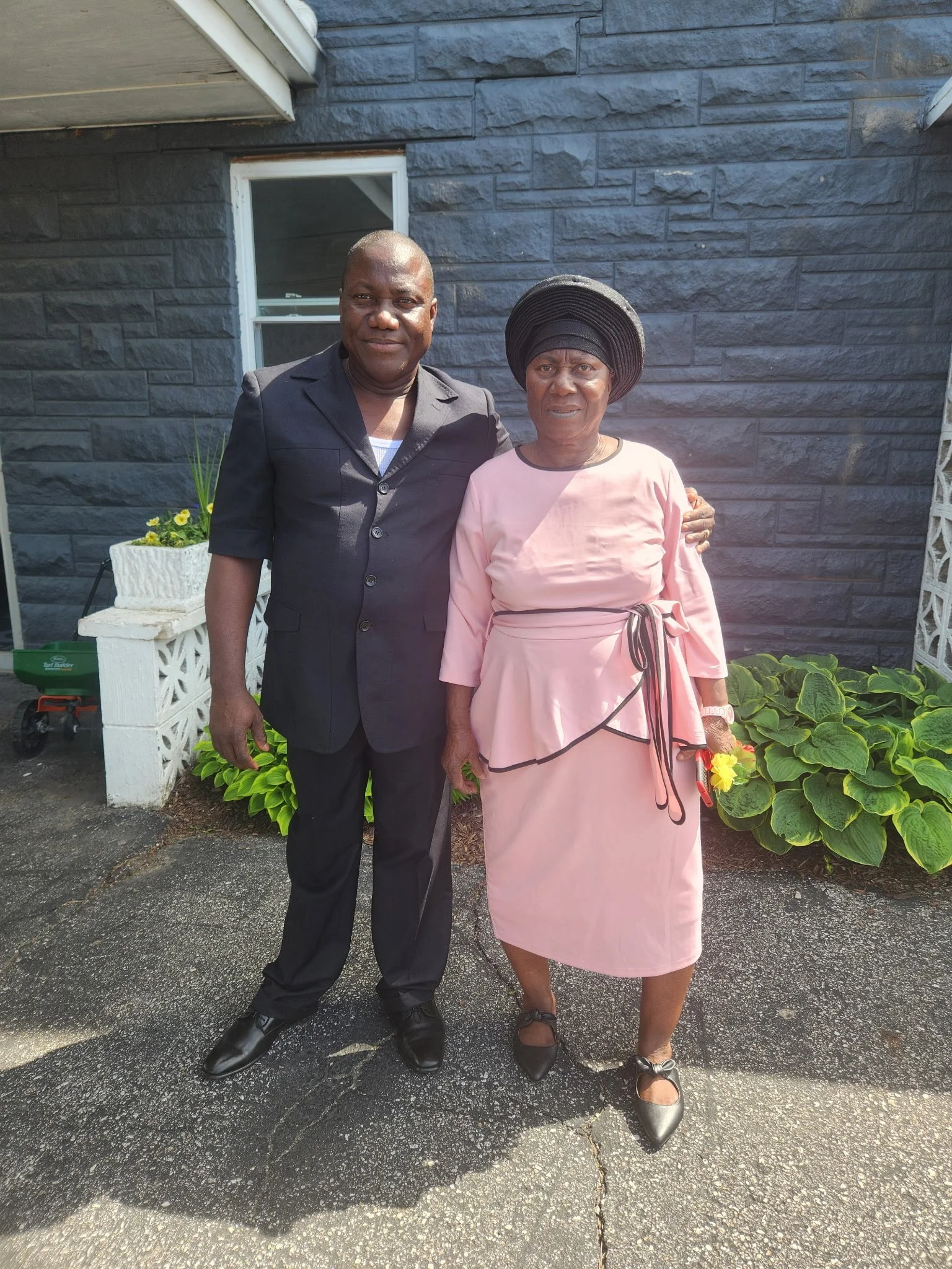 A man and a woman are standing outdoors on an asphalt surface, posing for a photo. The man is wearing a black blazer, black pants, and black shoes. The woman is in a pale pink dress with black trim, a bows at the waist, and a large black hat. They ar