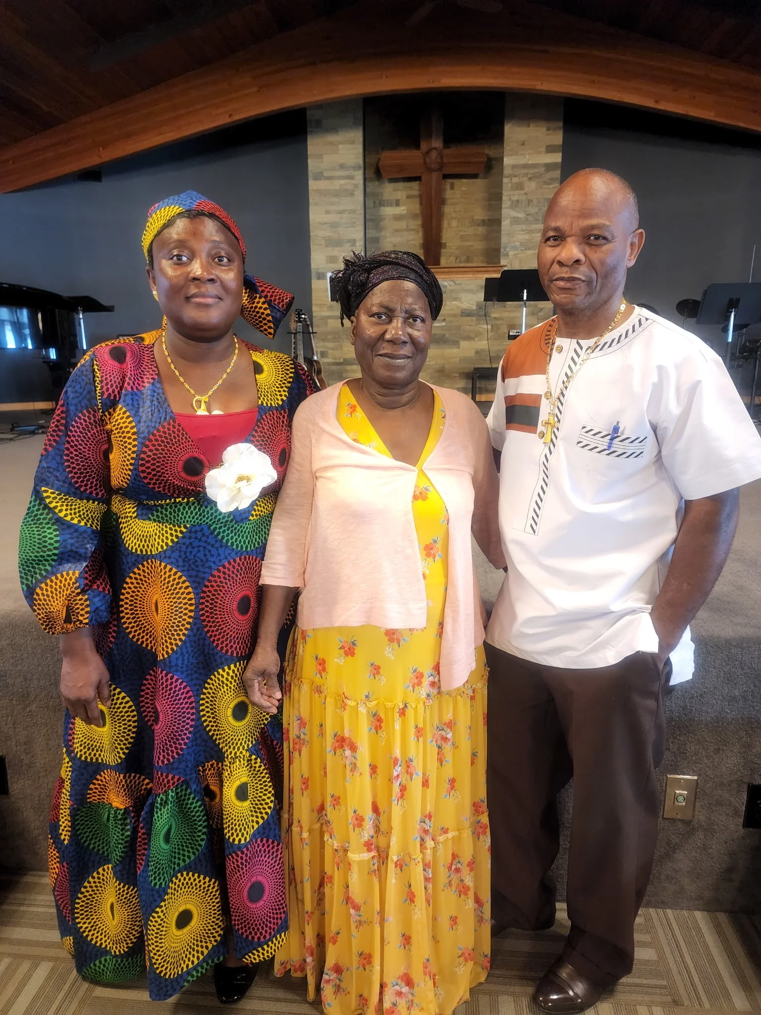 Three people standing inside a church, with a wooden cross on the wall behind them. The woman on the left is wearing a colorful patterned dress and headscarf, the woman in the middle is wearing a yellow floral dress with a light pink cardigan, and th