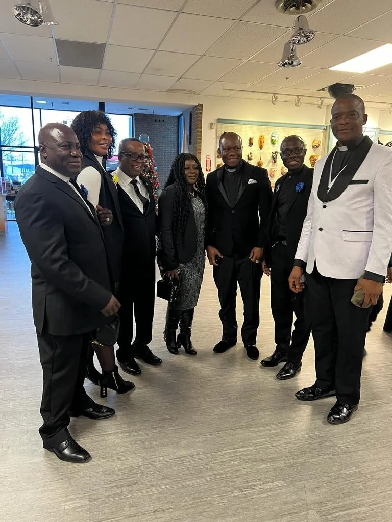Group of seven people dressed in formal attire standing together indoors, with Christmas decorations and window in the background.