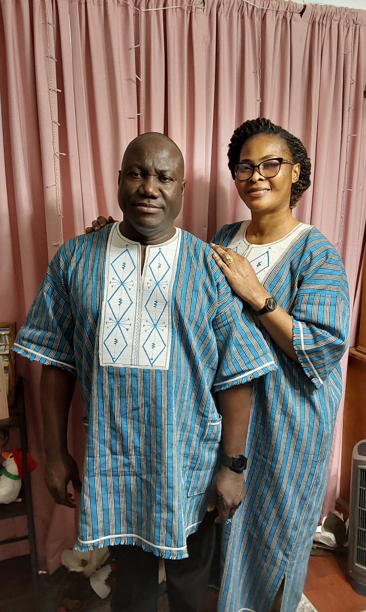 A man and woman wearing matching blue traditional African outfits, standing in front of pink curtains. The woman has glasses, a ring, and a watch, and the man has a watch. They are smiling and standing close together.