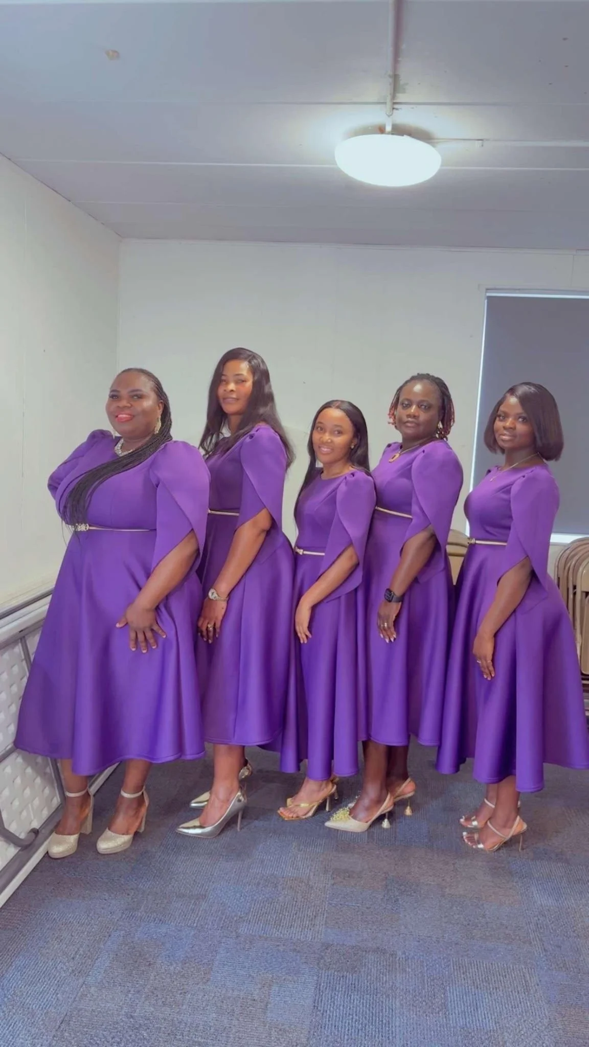 Five women standing in a line, wearing matching purple dresses and high heels in a room with white walls, grey carpet, and a window with a partially pulled down roller shade.
