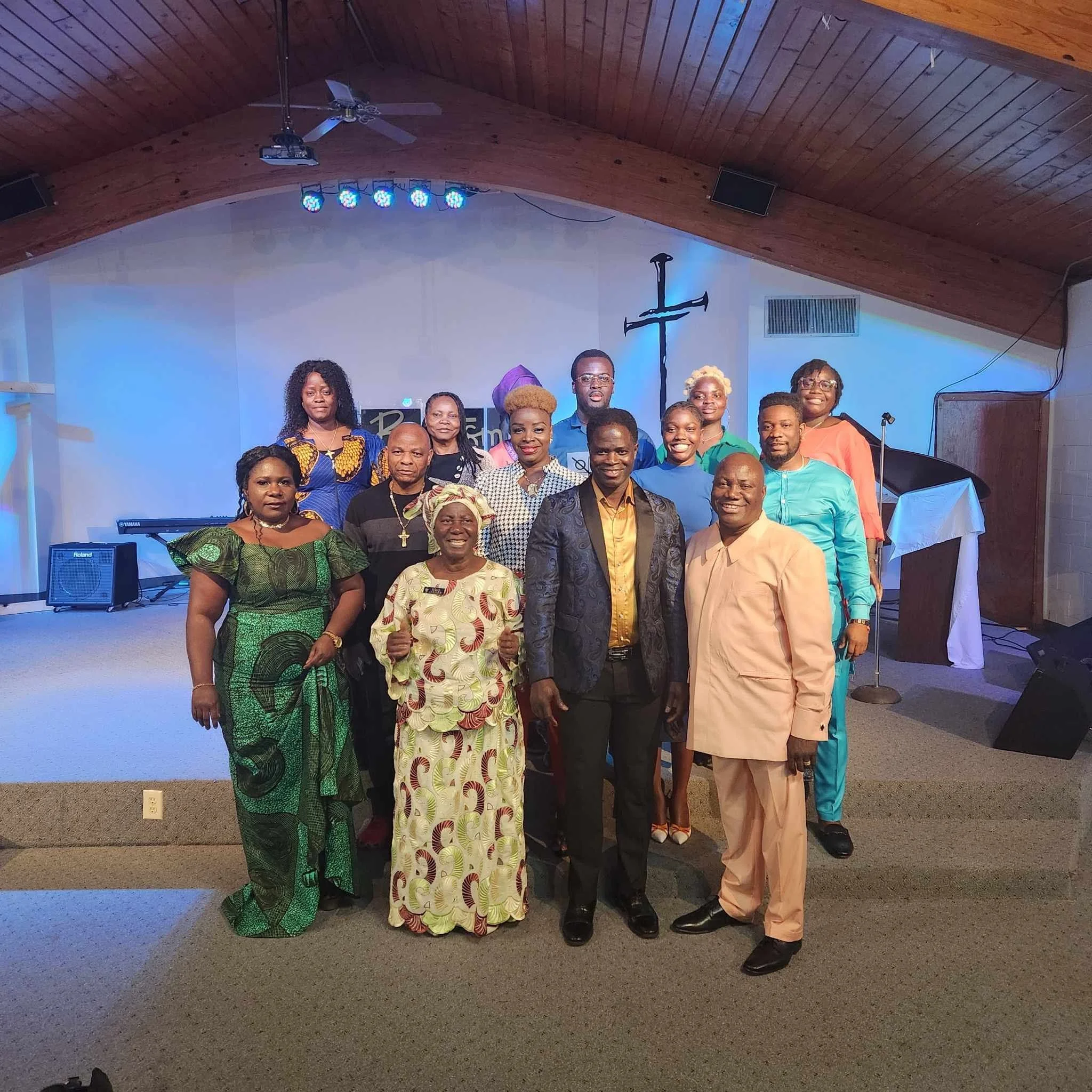 A group of diverse people standing together indoors in front of a white wall with a crucifix and stage lighting, smiling at the camera.
