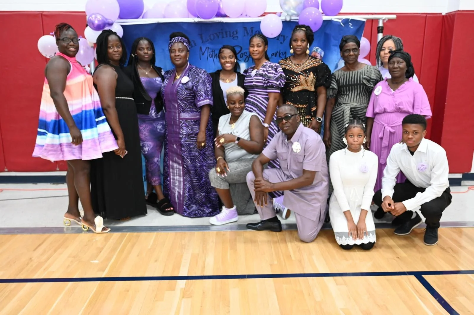 Group photo of diverse women and children in colorful attire at a celebration in a gymnasium with purple balloons and a blue banner in the background.