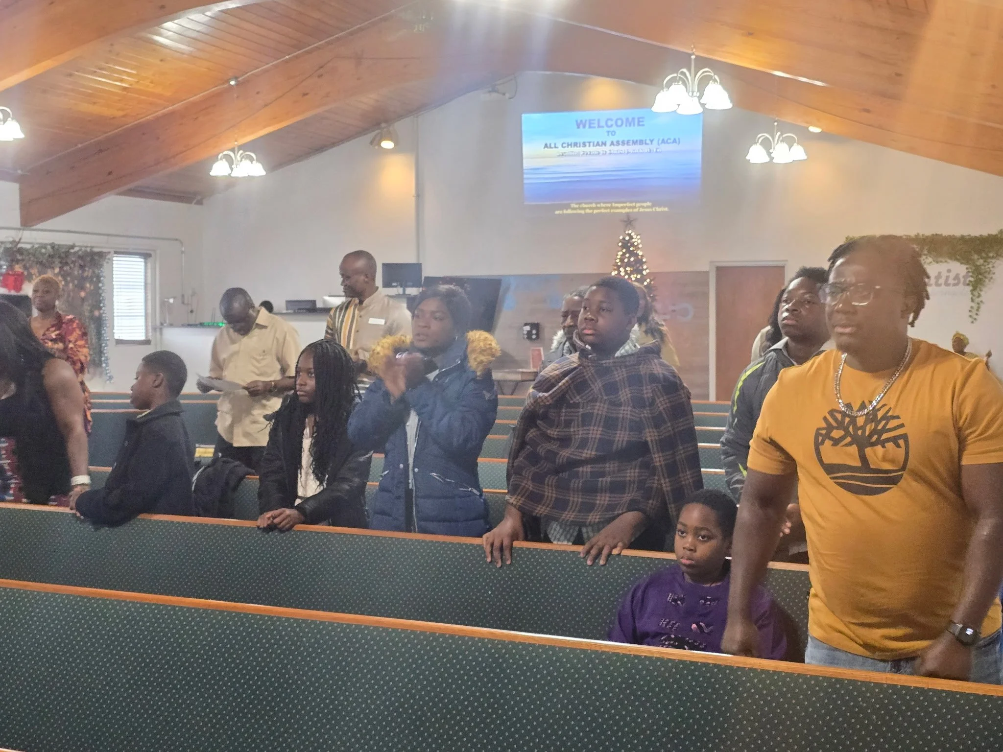 People gathered inside a church during a service, some standing and listening, with a wooden ceiling and a projection screen displaying a welcome message.