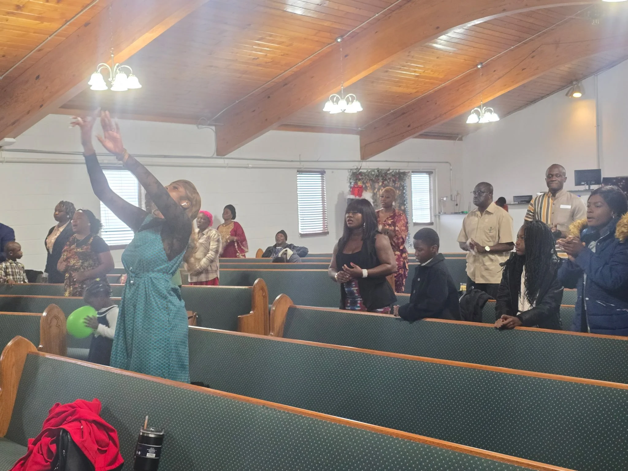 People gathered in a church service, some standing and some sitting, with a woman reaching up and touching the ceiling. The church has a wooden ceiling with hanging lights, and stained glass windows are visible in the background.