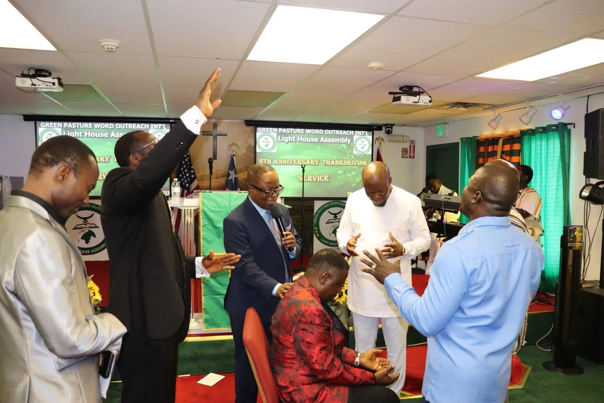 A group of people gathered at a church event, with some praying and one person raising his hand. There are banners and flags in the background. The event is celebrating the 5th anniversary of Green Pasture Word Outreach International, Light House Ass