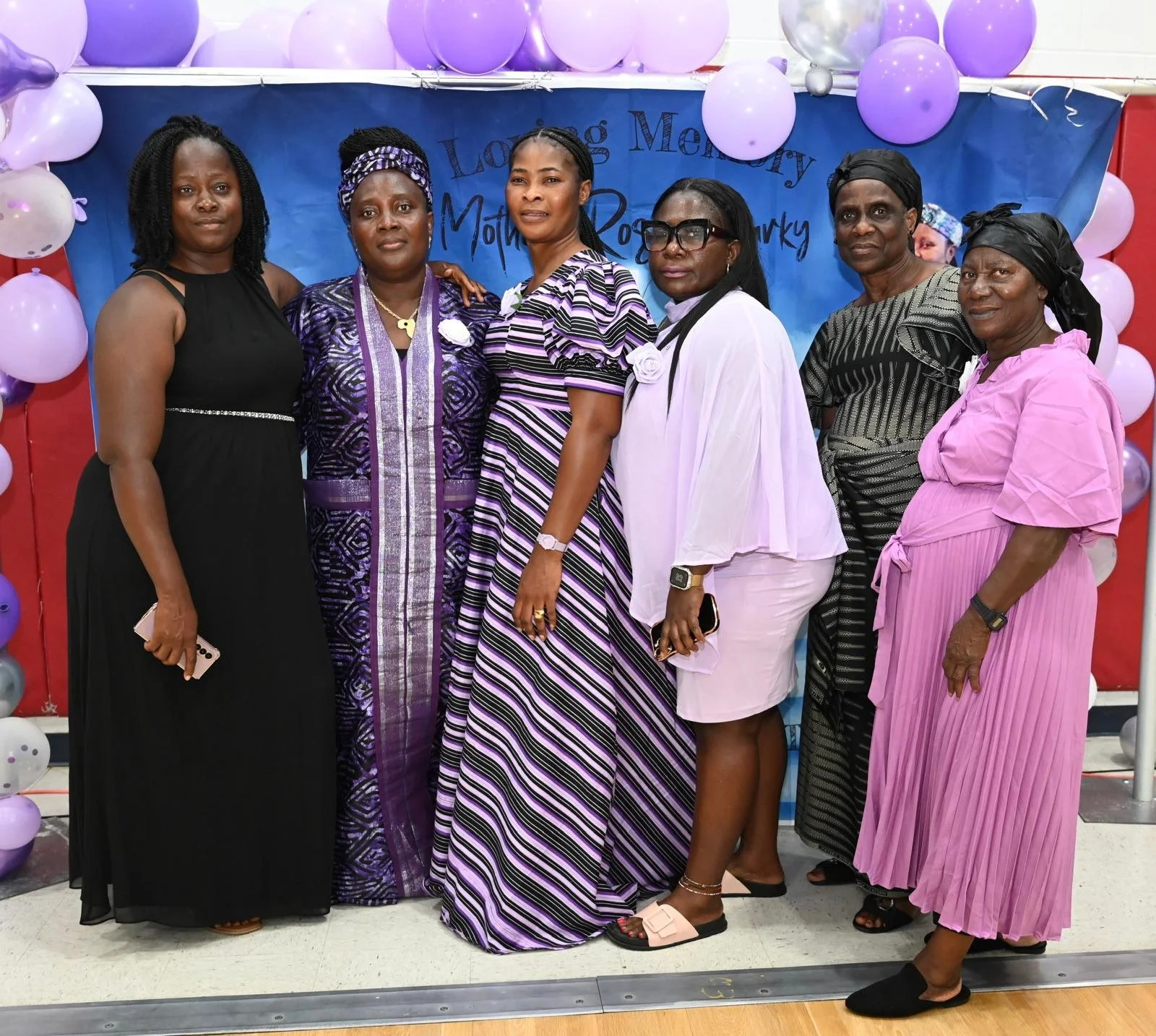 Group of seven women standing in front of a blue backdrop decorated with purple and white balloons. The women are dressed in various shades of purple, black, white, and pink, and some are wearing head wraps or glasses.