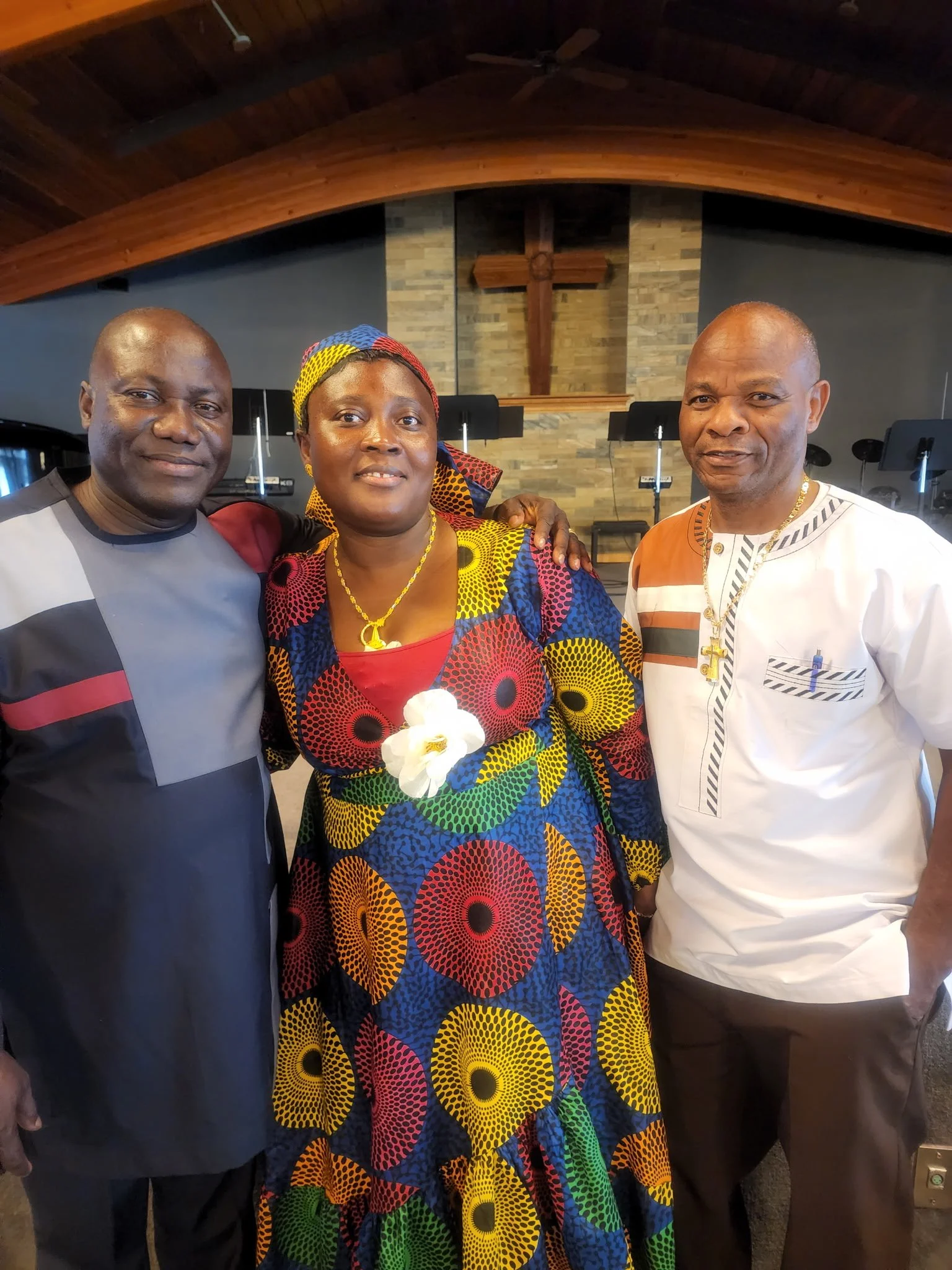 Three people, two men and one woman, posing together inside a church with a wooden cross on the wall behind them. The woman is wearing a colorful dress and headscarf, while the men are dressed in casual shirts.