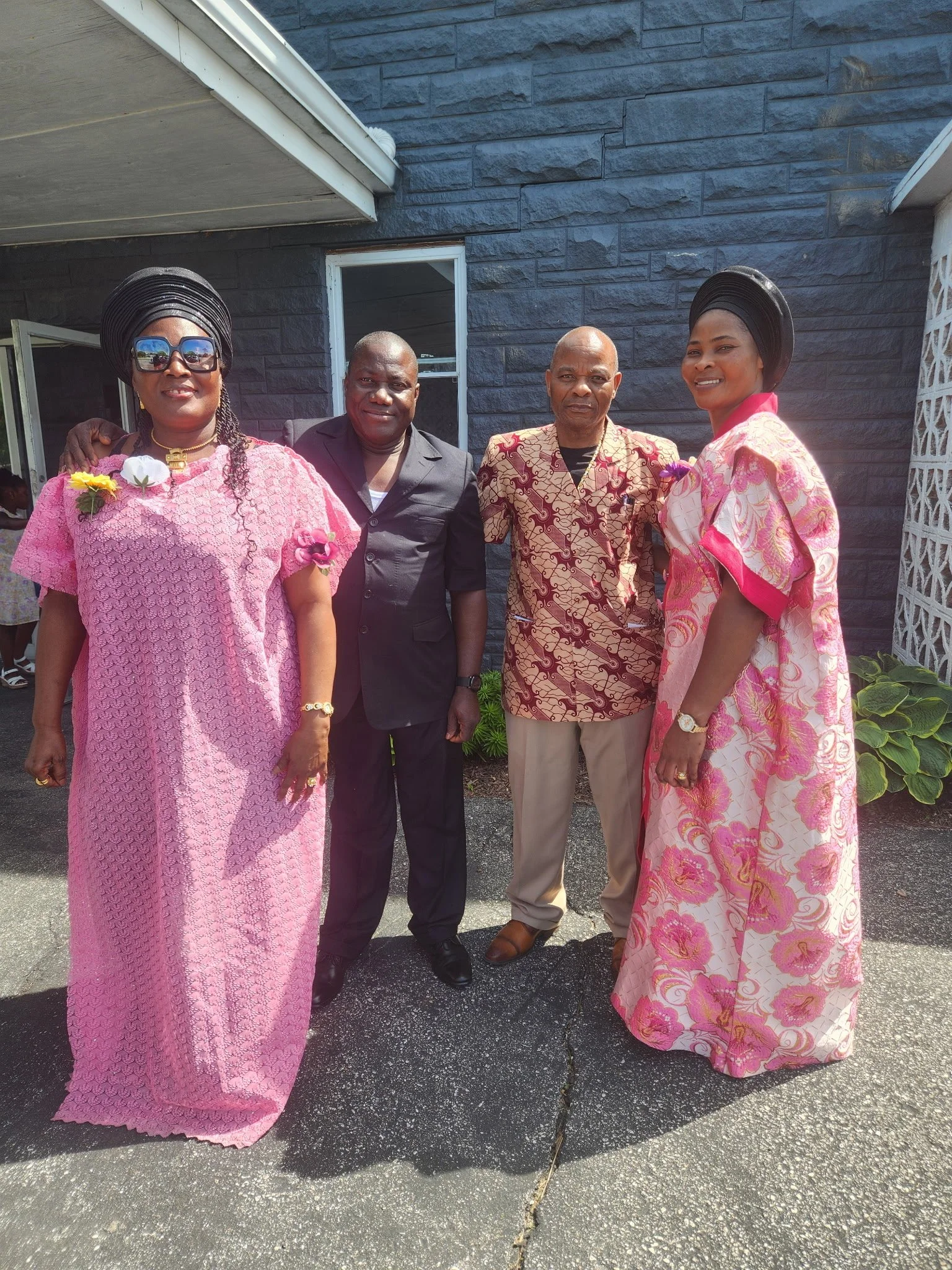 Four individuals standing outside in front of a dark brick building, two women dressed in traditional pink dresses and two men in dark suits, all smiling.