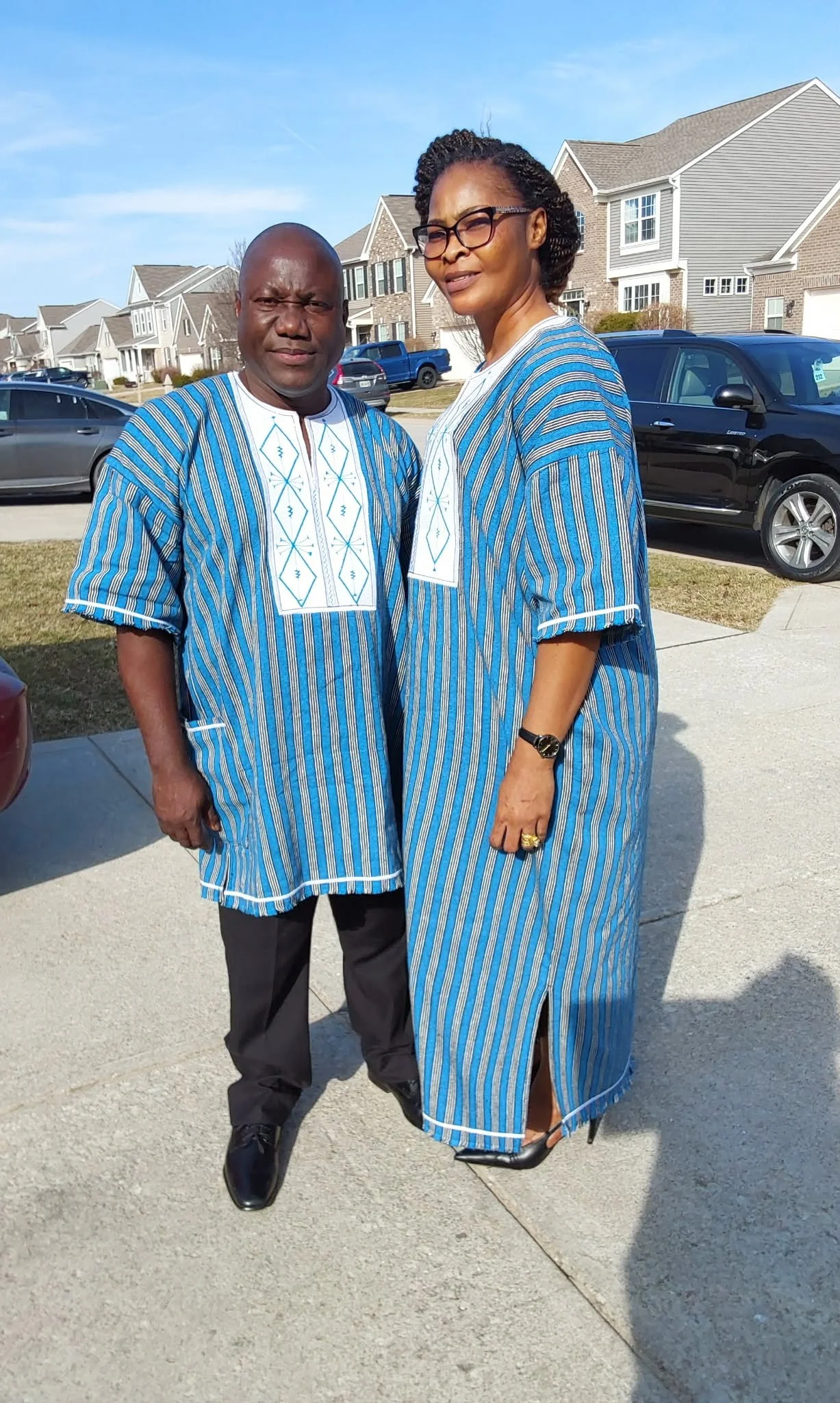 A man and a woman standing outside on a suburban street, wearing matching blue and white striped traditional clothing. The man is bald and wearing glasses, while the woman has short curly hair and is wearing a watch and rings. There are houses and ca
