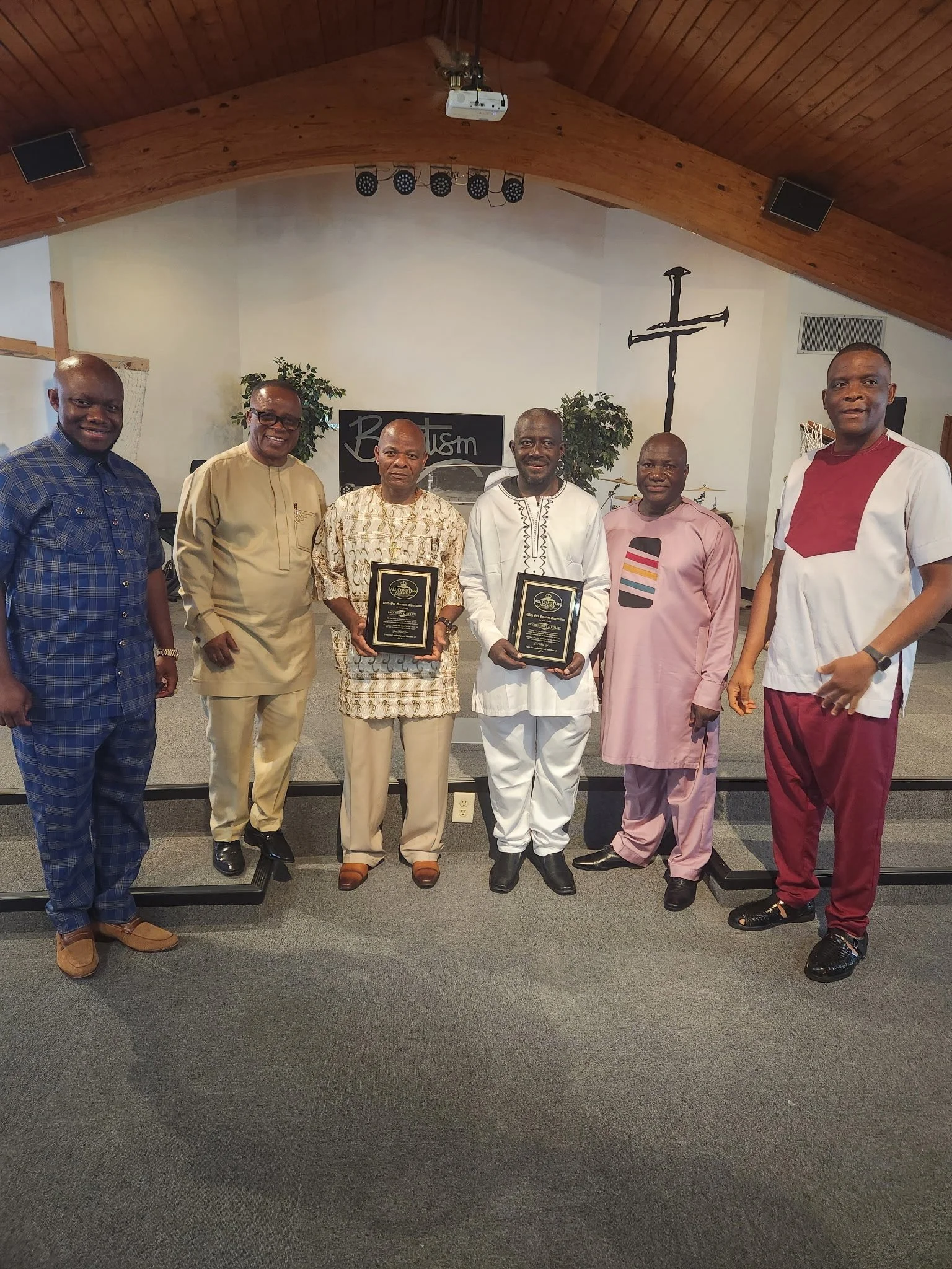 Six men standing inside a church, with three holding plaques. The background features a cross, framed by two potted plants, a sign that says 'Redemption,' and church equipment. The church has a vaulted wooden ceiling.