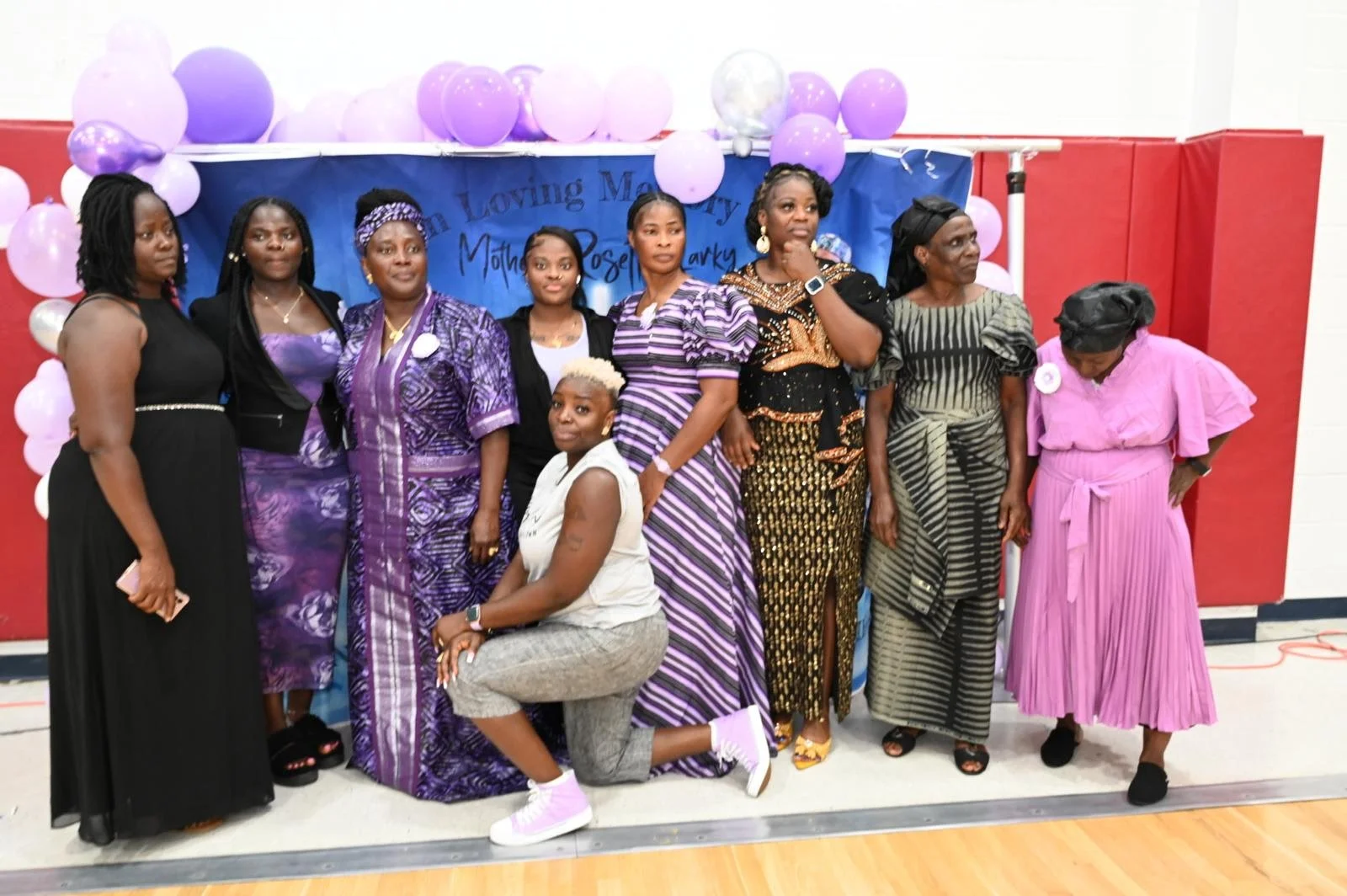 Group of women in colorful dresses, gathered for a celebration, with purple and white balloons and a blue backdrop.
