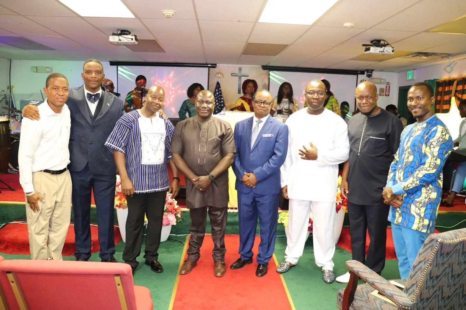 Group of nine men standing on a red carpet in a church, with a cross and musical instruments in the background.