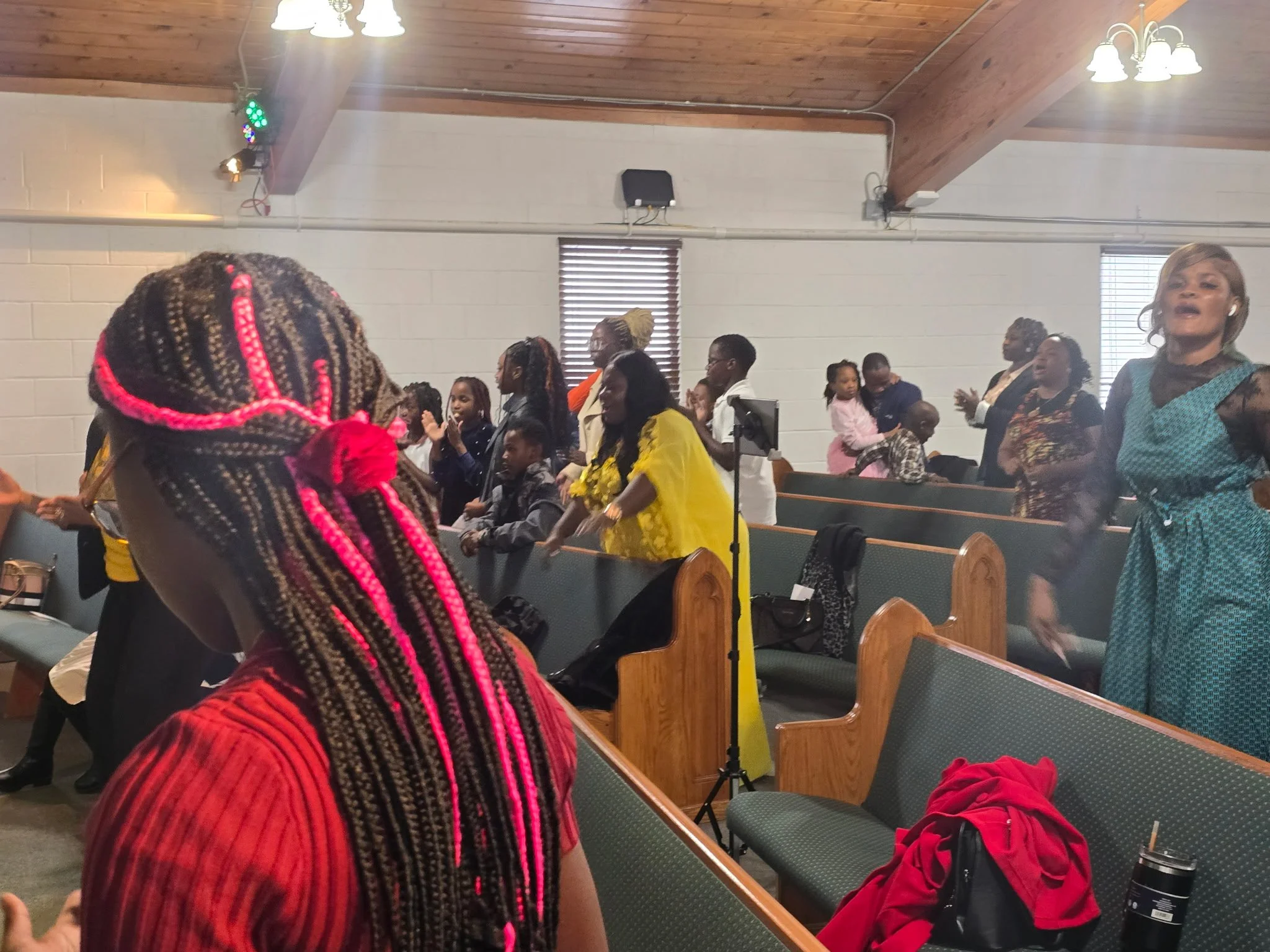 People gathered in a room with wooden ceiling, some standing and some sitting, in what appears to be a church or community hall. There are wooden pews, window blinds, and various individuals, some clapping or engaged in activity.