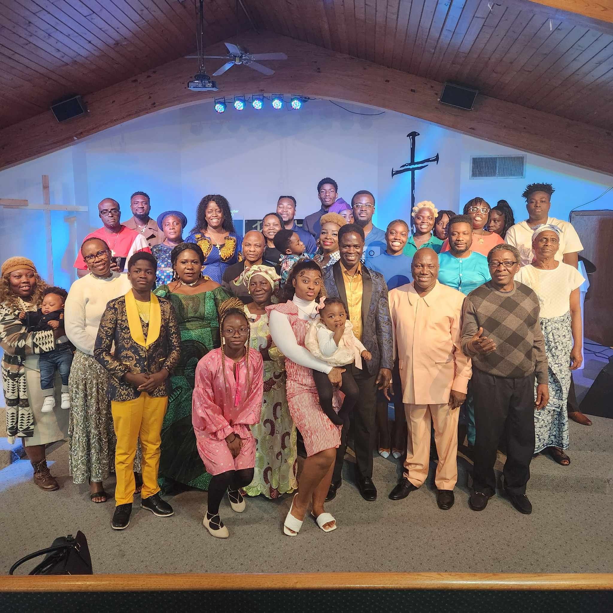 A large group of diverse people gathered indoors for a photo on a stage. The background features a white wall with a black cross symbol, a ceiling fan, and several stage lights. The group includes men, women, and children dressed in colorful clothing