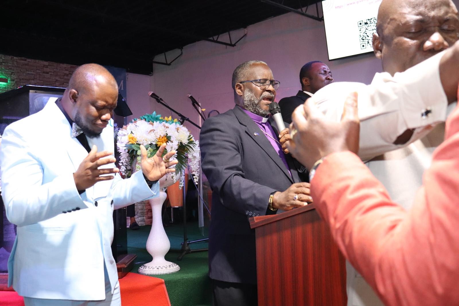 A group of men in formal attire praying at a church service, with one man speaking into a microphone and others with eyes closed in prayer.