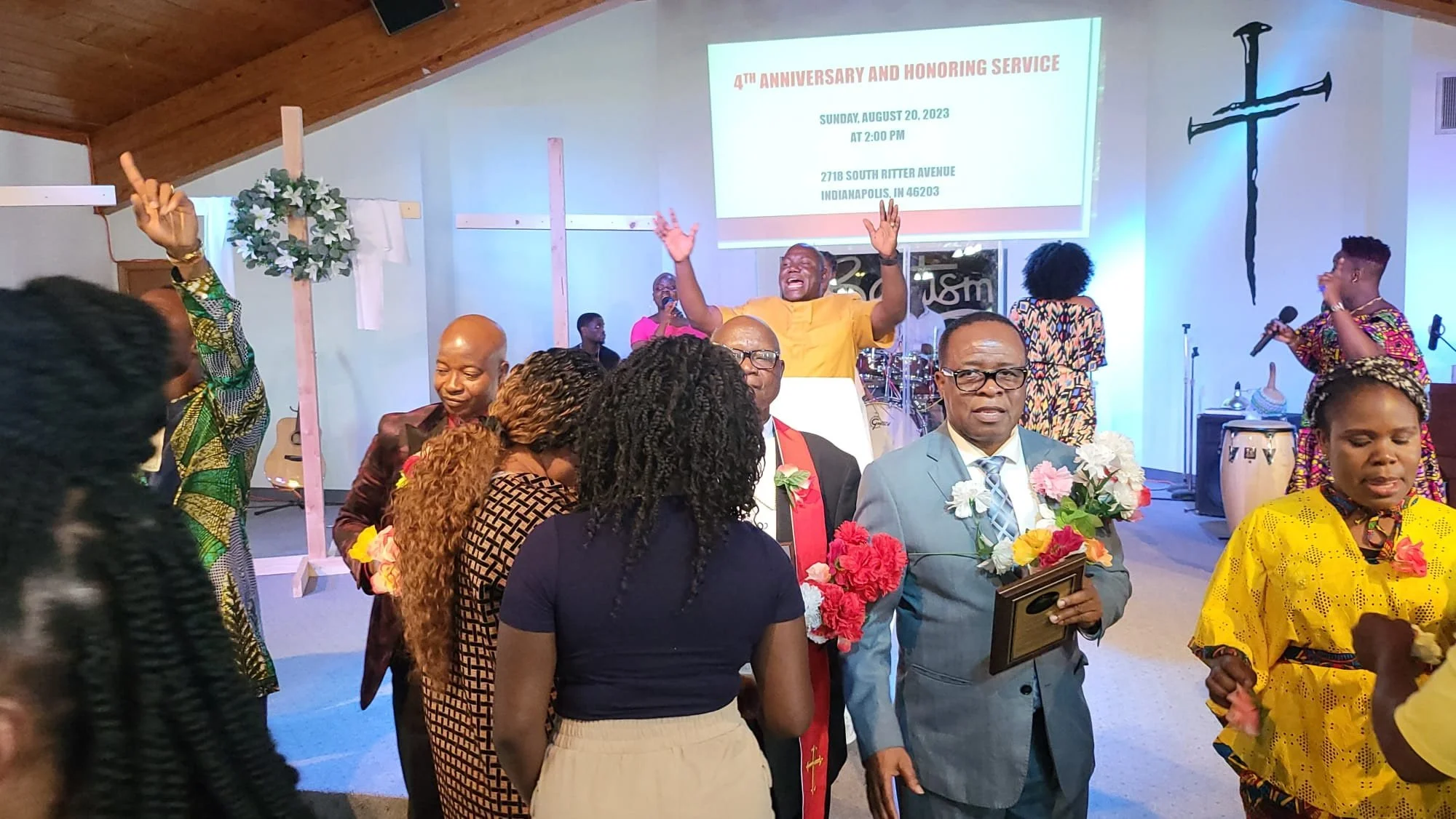 A church service celebrating its 4th anniversary, held on August 20, 2023, at 2:00 PM at 2718 South Ritter Avenue, Indianapolis, Indiana. The image shows a congregation of men and women, some holding framed certificates and flowers, with a speaker at