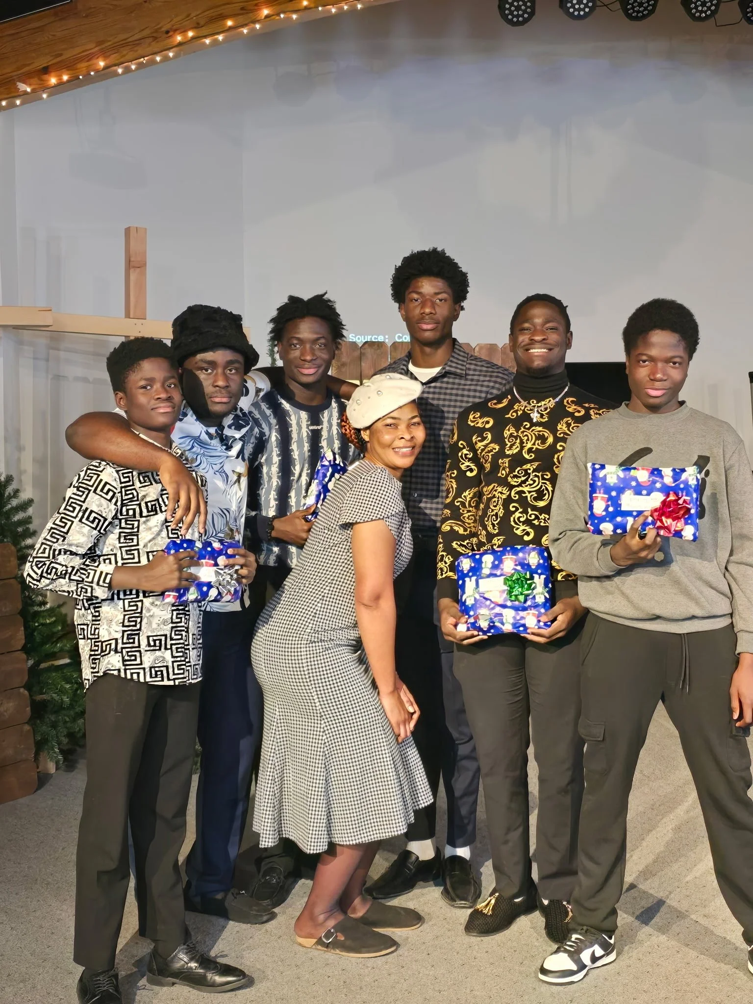A group of nine people, standing closely together and smiling, holding wrapped presents. The group includes both males and females, with some individuals dressed in patterned and casual clothing. The background features a decorated space with string 