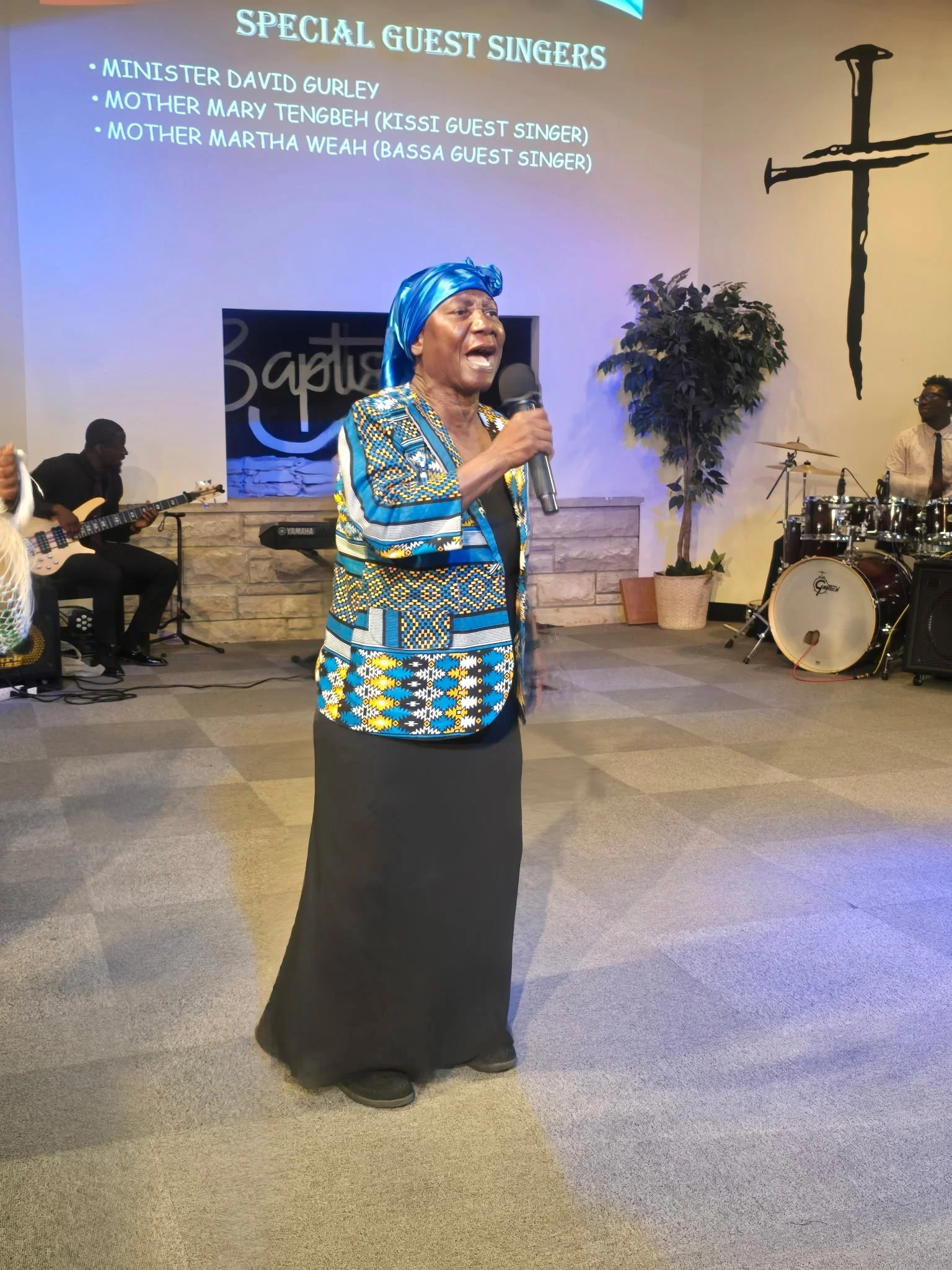 A woman wearing a colorful patterned dress and a blue headscarf sings into a microphone on stage. Behind her, a band with a guitar, keyboard, and drums performs. A large screen displays a list of special guest singers, including Minister David Gurley
