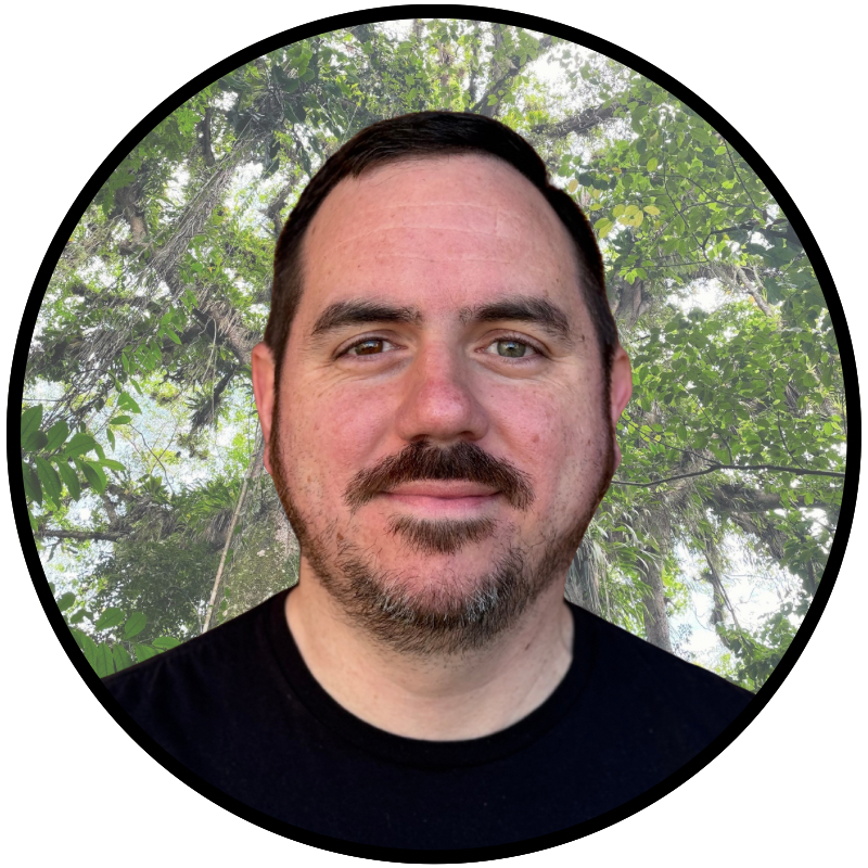 A man with dark hair and a beard, smiling, with a lush green forest background.