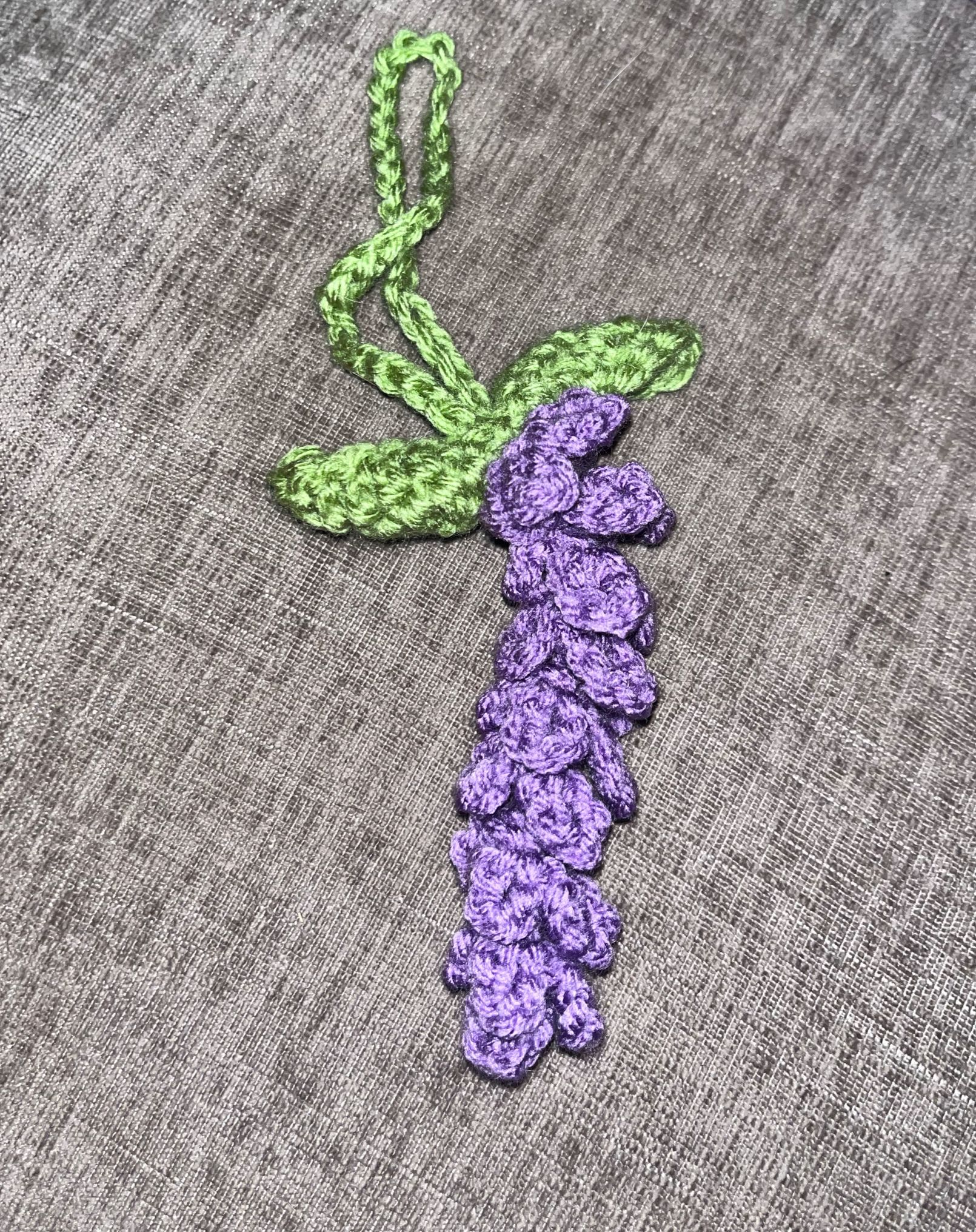 A handmade embroidered lavender flower with green leaves and a green stem on a gray fabric background.
