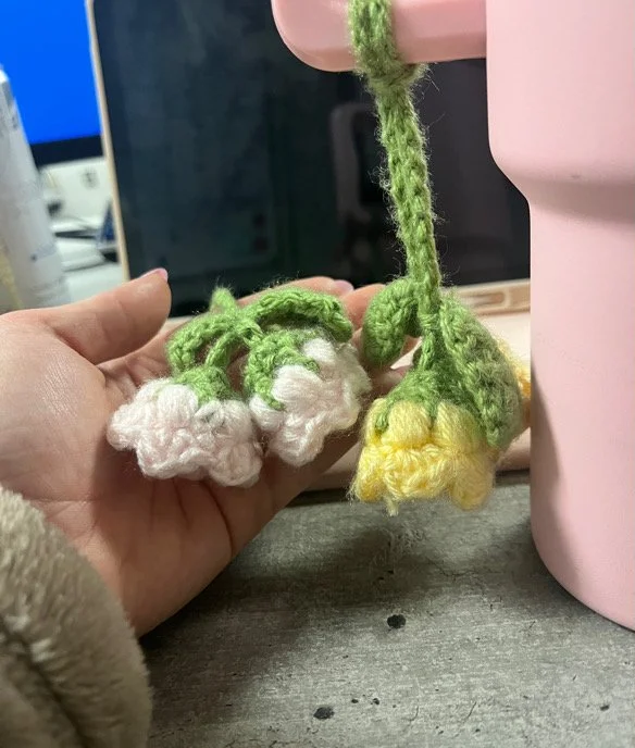 Hand holding two small crocheted flowers, one white with green stems and leaves, the other yellow with green stems, next to a pink object on a desk.