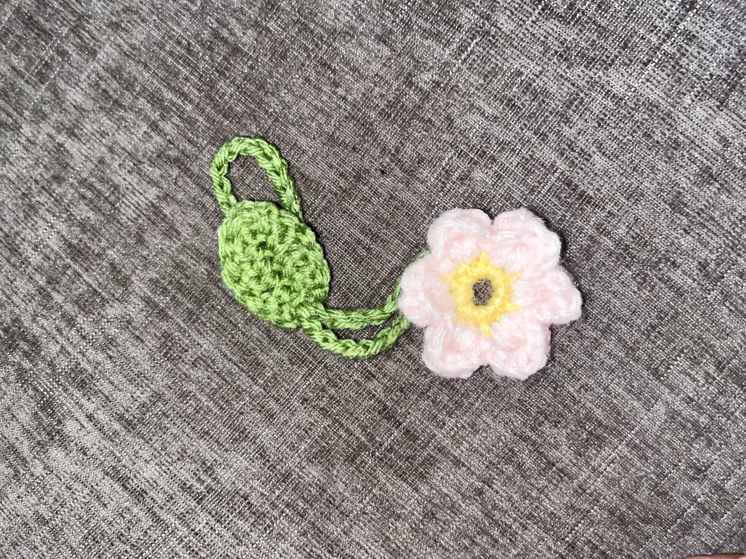 Crochet floral patch with pink flower and green leaf on gray fabric background.