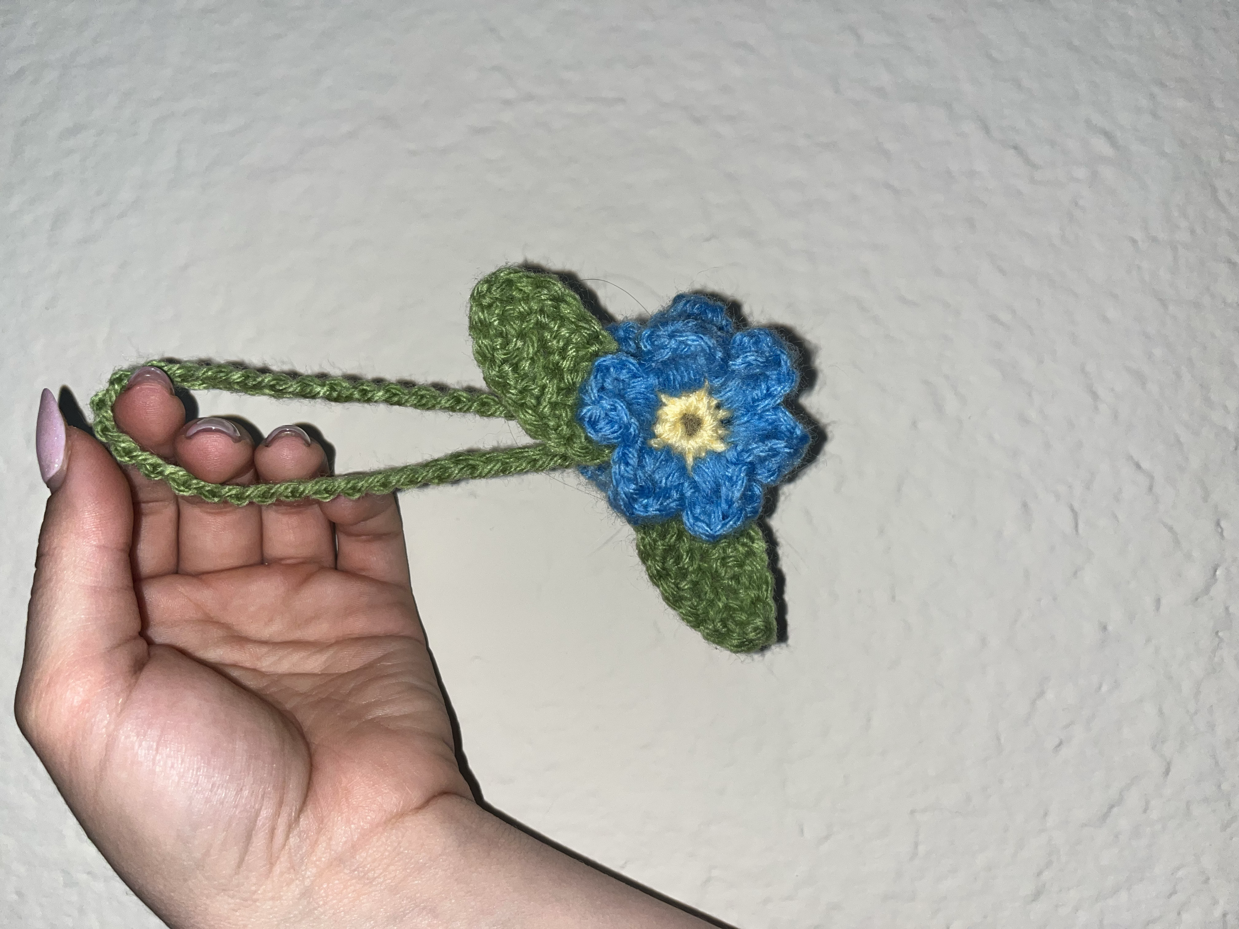 Hand holding a crocheted blue flower with green leaves against a white wall.
