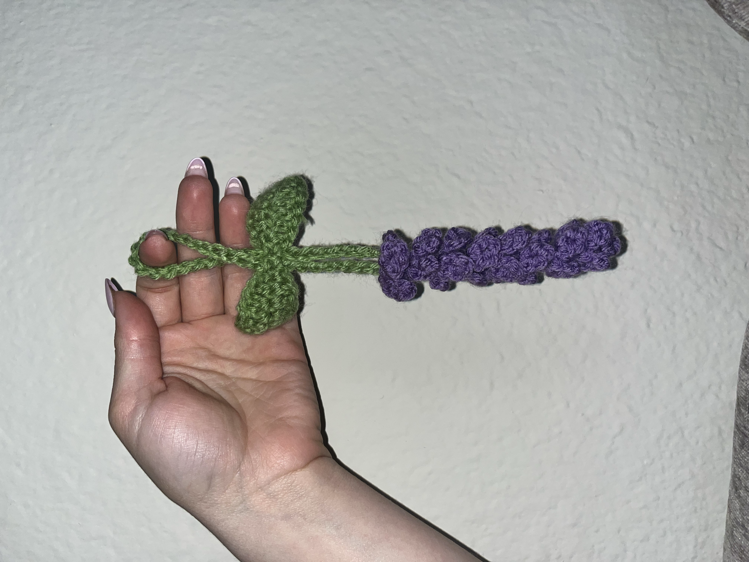 Hand holding a crocheted lavender and green dragonfly with purple body and green wings against a plain wall background.