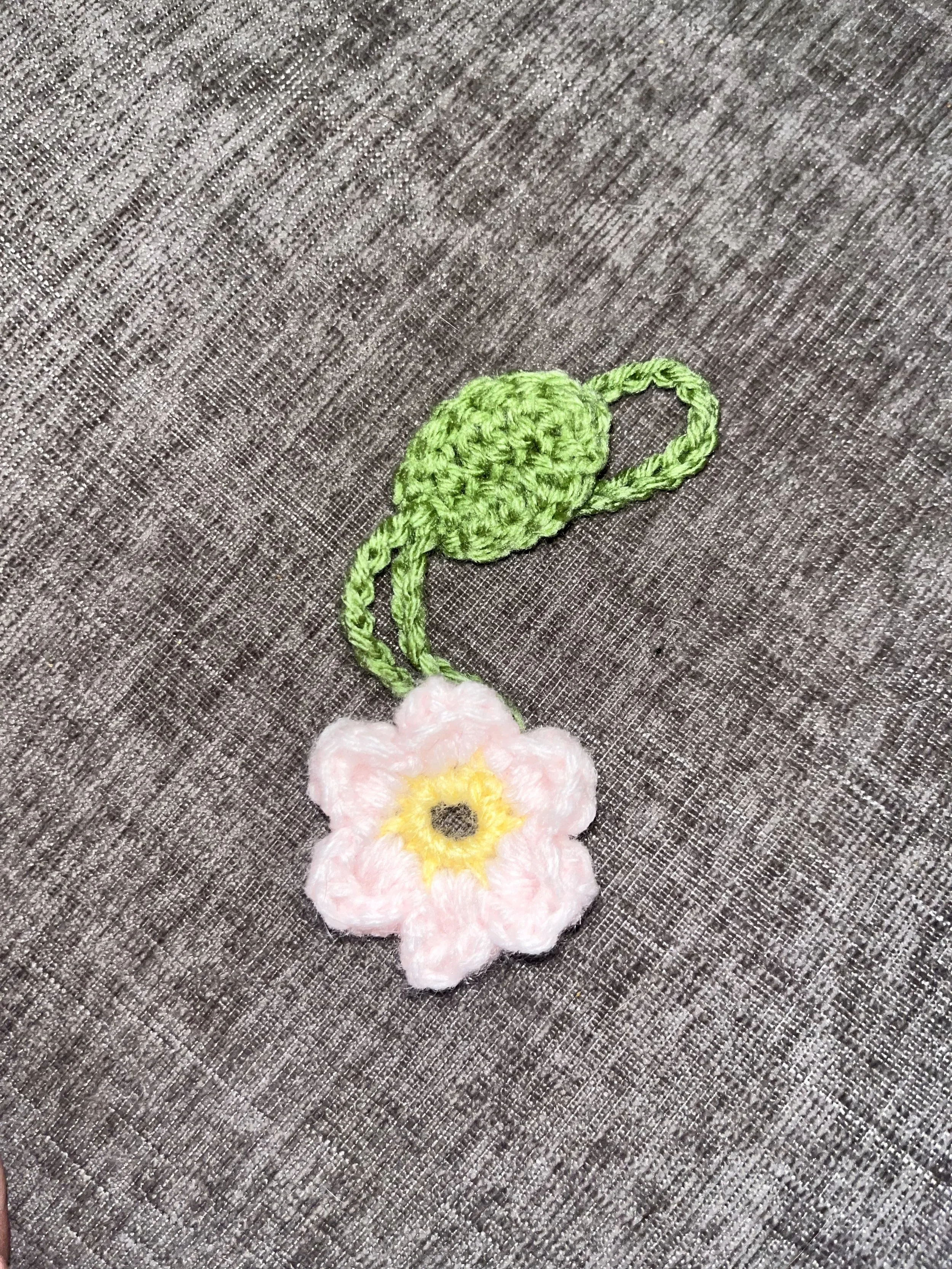Handmade crochet flower with pink petals, yellow center, green leaves, and stem on grey fabric background.