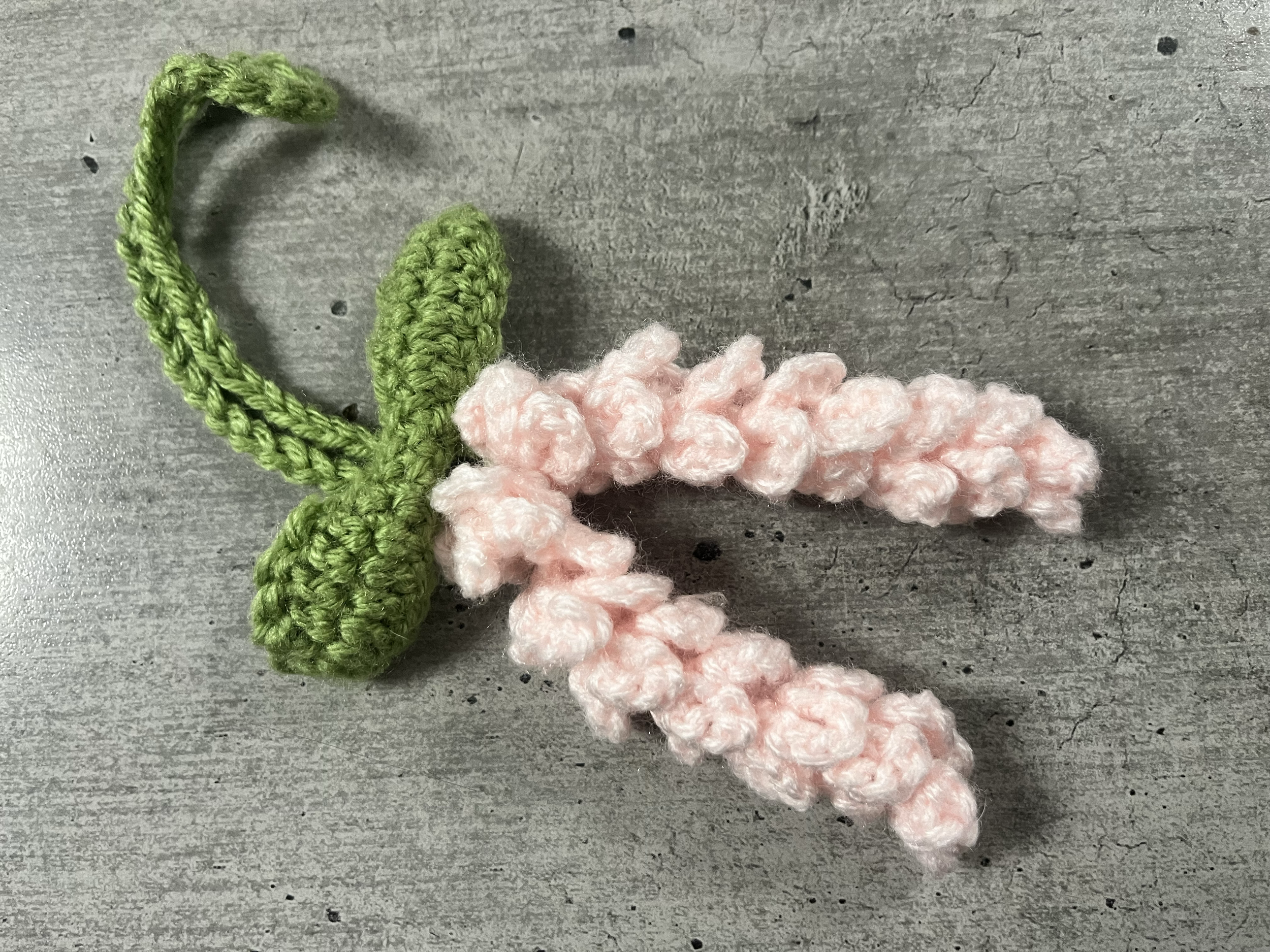 A small, pink and green, hand-crocheted flower with pink petals and green stem and leaves, lying on a gray surface.