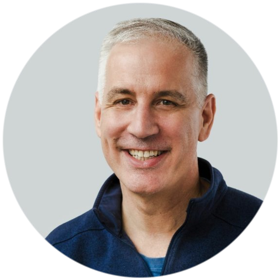 A smiling middle-aged man with short gray hair, wearing a navy blue zip-up jacket, in front of a light gray background.