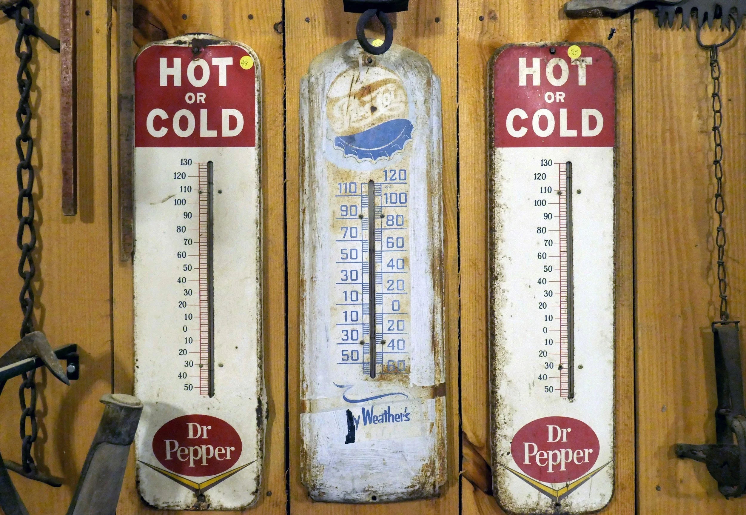 Three vintage old-fashioned thermometers mounted on a wooden wall, two labeled "HOT or COLD" with a red top and white bottom, and one in the middle with a blue and yellow design. The thermometers have rust and wear, indicating age. There are tool hooks on either side, with tools hanging from chains.