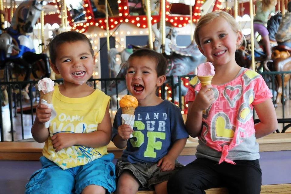 Three young children sitting together, each holding an ice cream cone and smiling at a carnival or amusement park with bright lights and a carousel in the background.