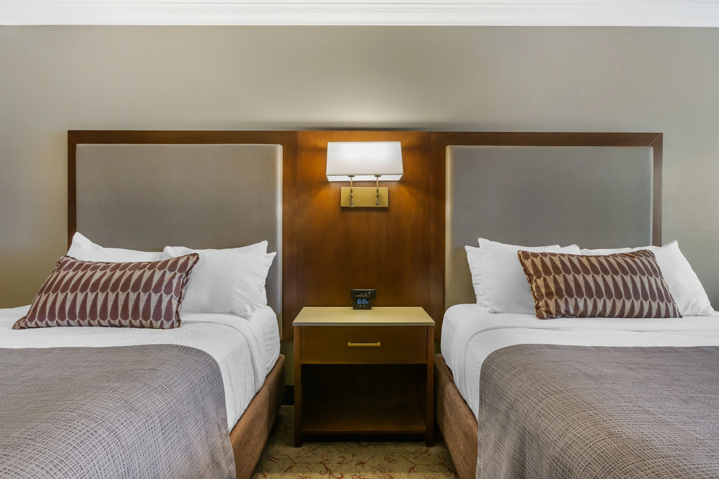 Two hotel beds with headboards, pillows, and bedspreads separated by a nightstand, with a wall-mounted lamp and clock on the wall.