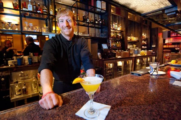 Smiling bartender standing behind a bar with a yellow cocktail glass in front of him, inside a well-lit bar or restaurant.