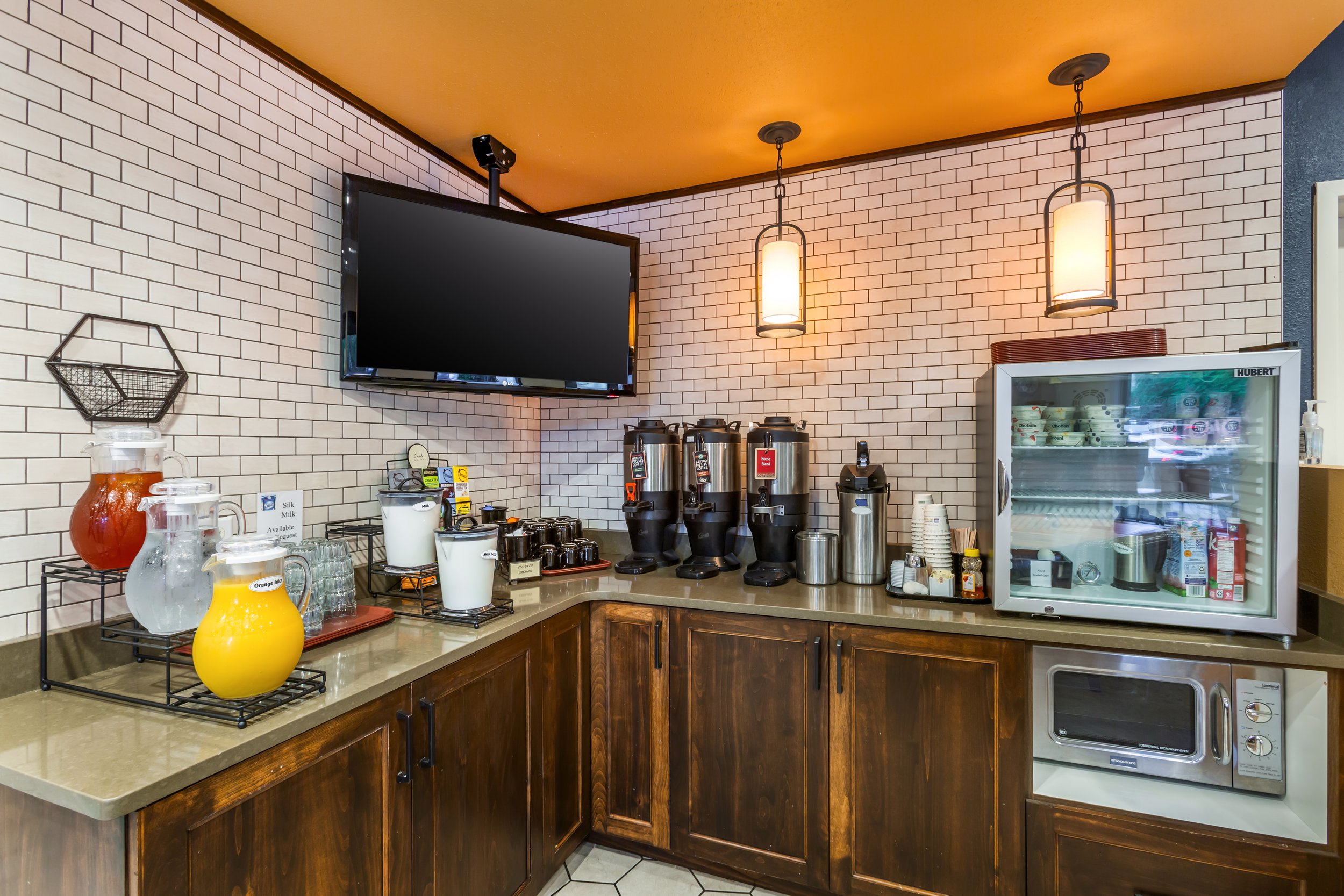 Coffee station with three coffee dispensers, a small refrigerator, a microwave, and various glass pitchers with fruit infusions, on a countertop with dark wooden cabinets.