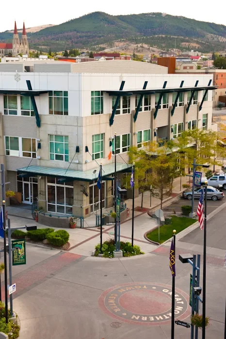 Modern multi-story building with glass windows and black structural supports, located on a city street with flags, trees, and parked cars, with hills and a church in the background.