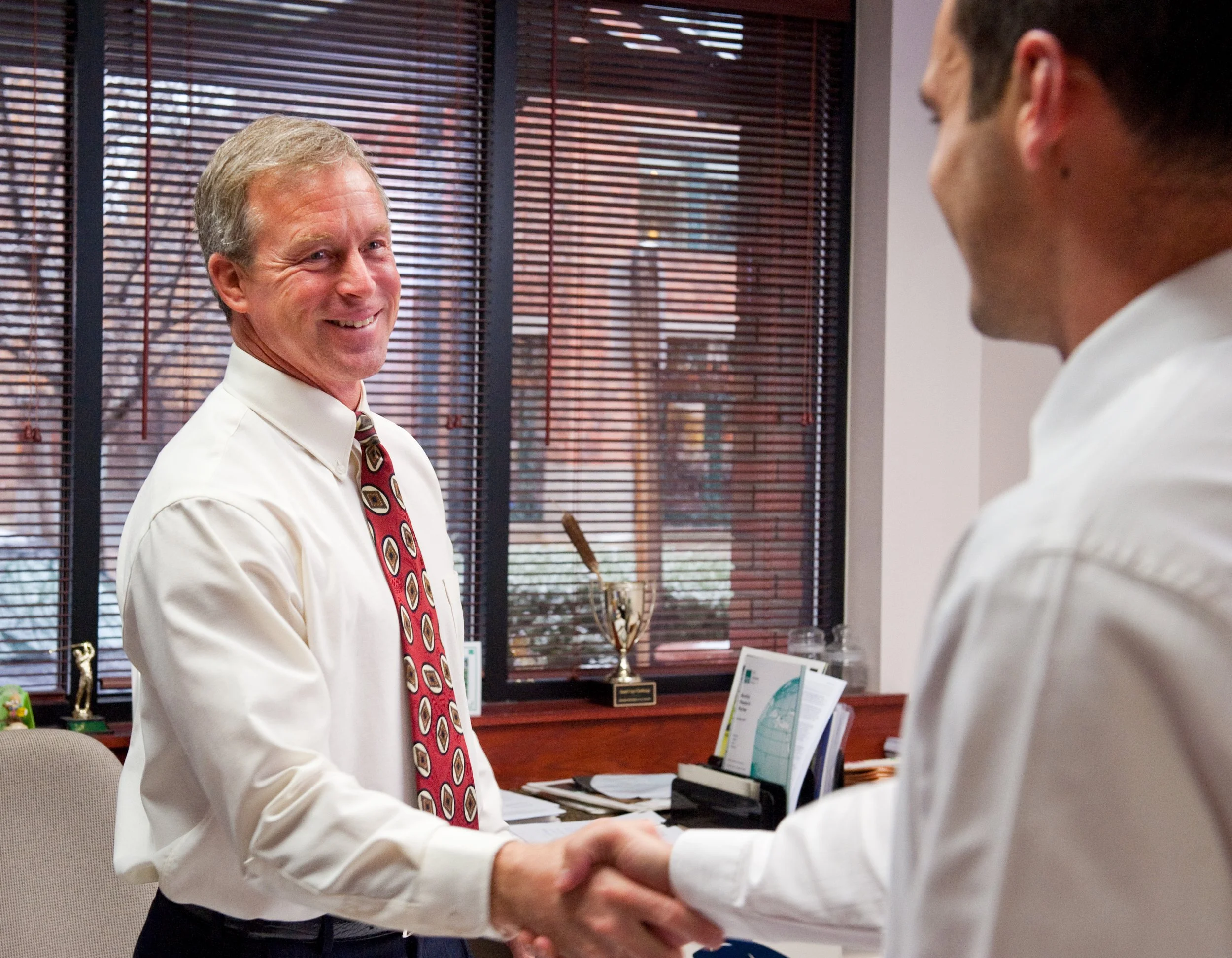 Two men shaking hands in an office, one smiling. The office has blinds, a trophy, and some documents on the desk.
