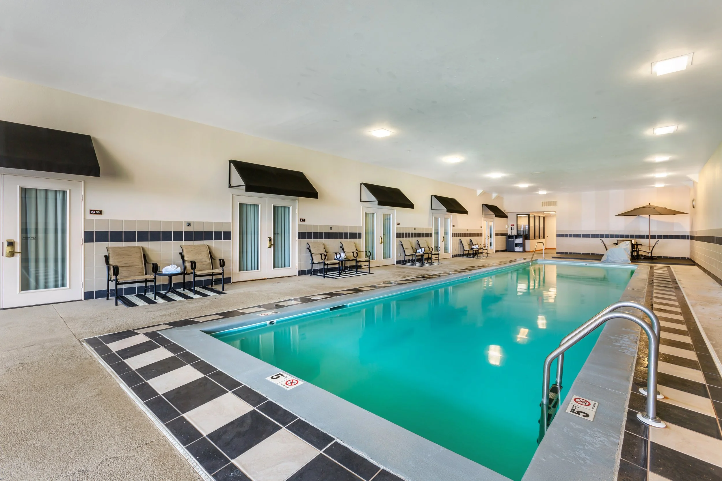 Indoor hotel swimming pool with chairs and umbrellas along the sides, black awnings above doors, and a tiled border around the pool.