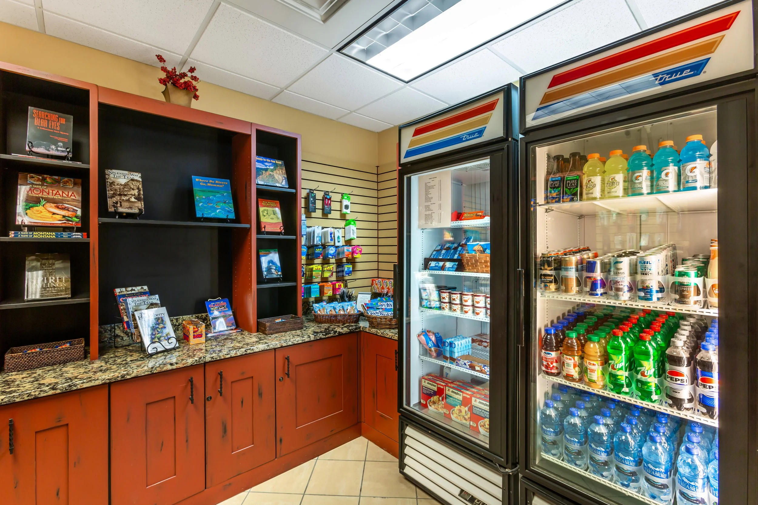 Inside a small store with a granite counter, black shelves with books, a cabinet, and a refrigerator stocked with drinks.