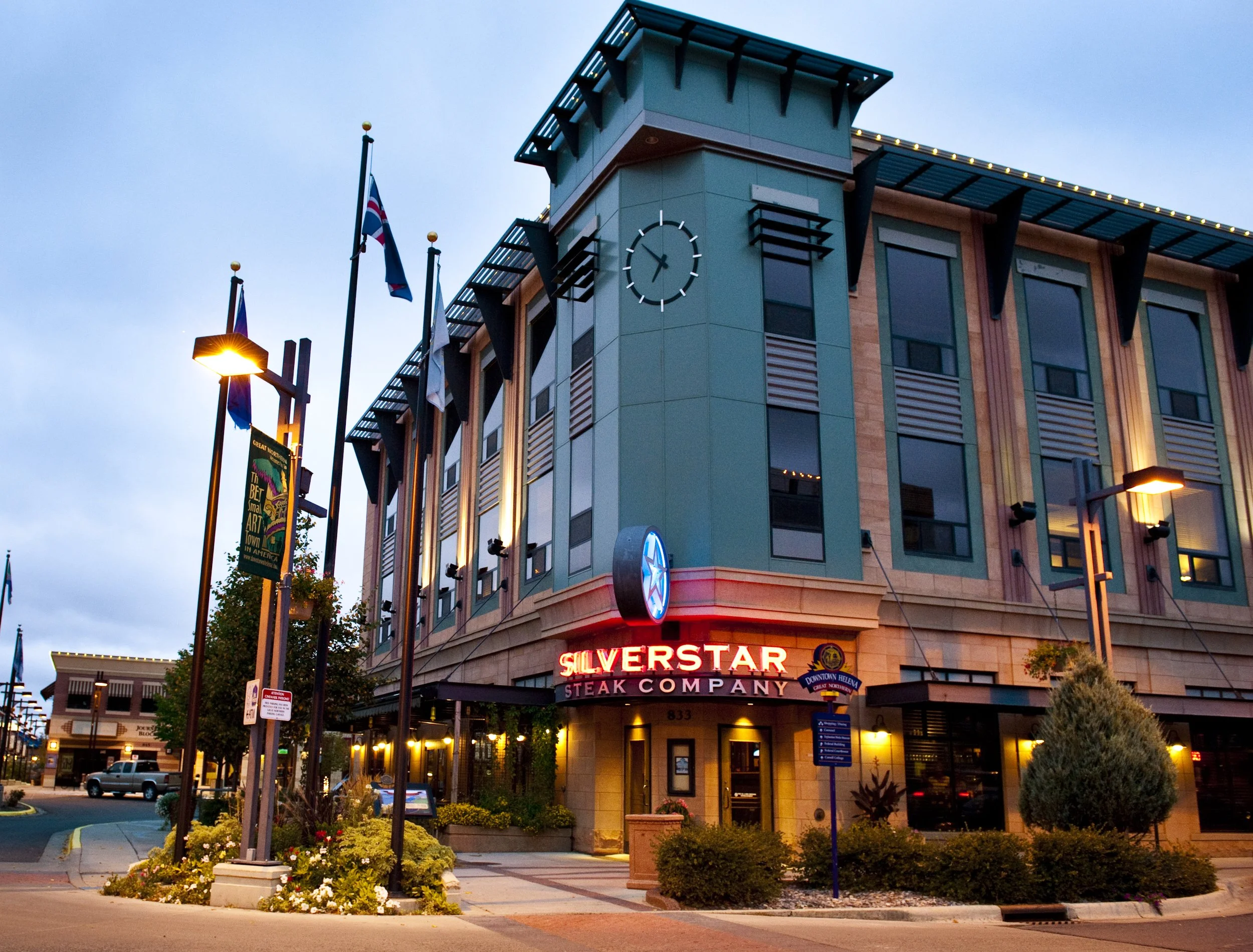 A multi-story building with a clock on top, illuminated signage reading 'SILVERSTAR STEAK COMPANY', and flags outside during early evening. The street is lined with lamp posts, trees, and flowerbeds.