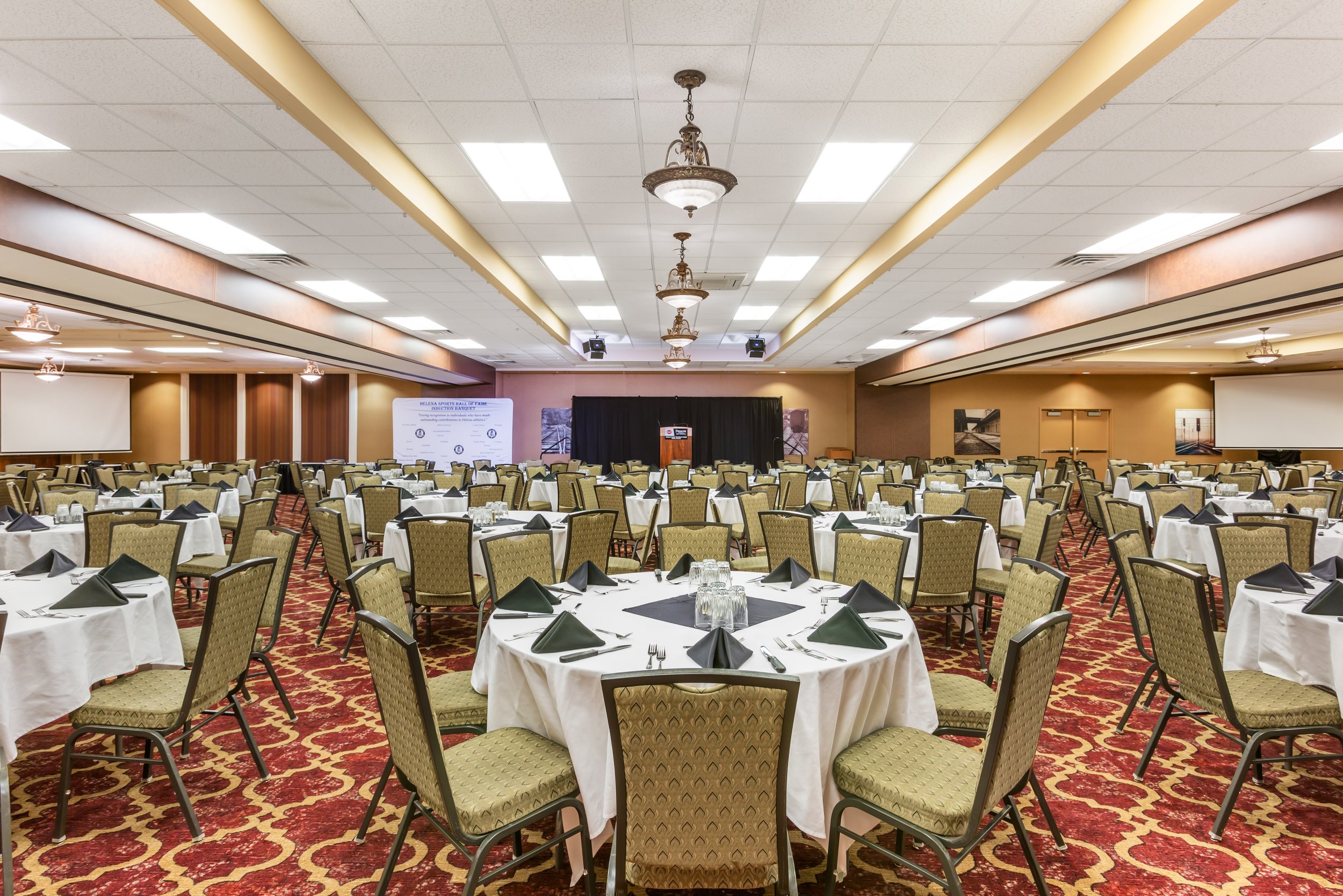 A large banquet room set up for a formal event with round tables covered in white tablecloths, black napkins, and glassware, with a stage at the front and decorative chandeliers hanging from the ceiling.