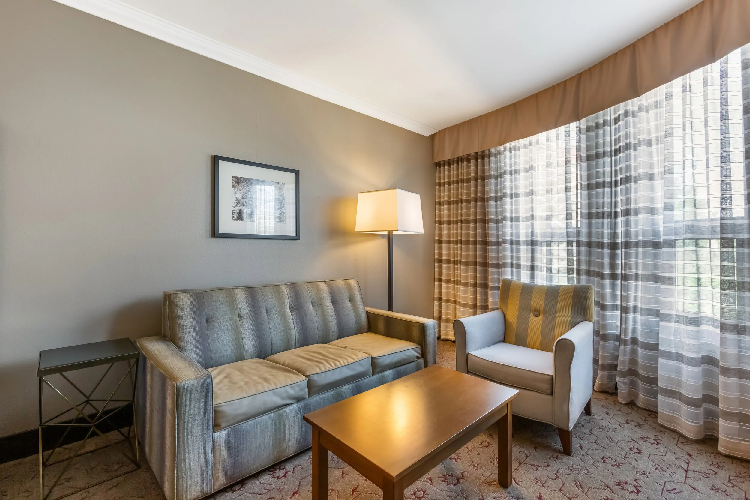 A living room with a beige wall, a gray striped sofa, a small black side table, a yellow and white armchair, a wooden coffee table, a floor lamp, and large windows with checkered curtains.