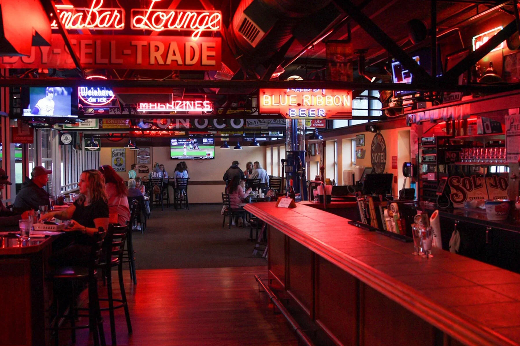 Interior of a sports bar with neon signs, TVs showing baseball and soccer games, wooden bar counter, and patrons sitting at tables.